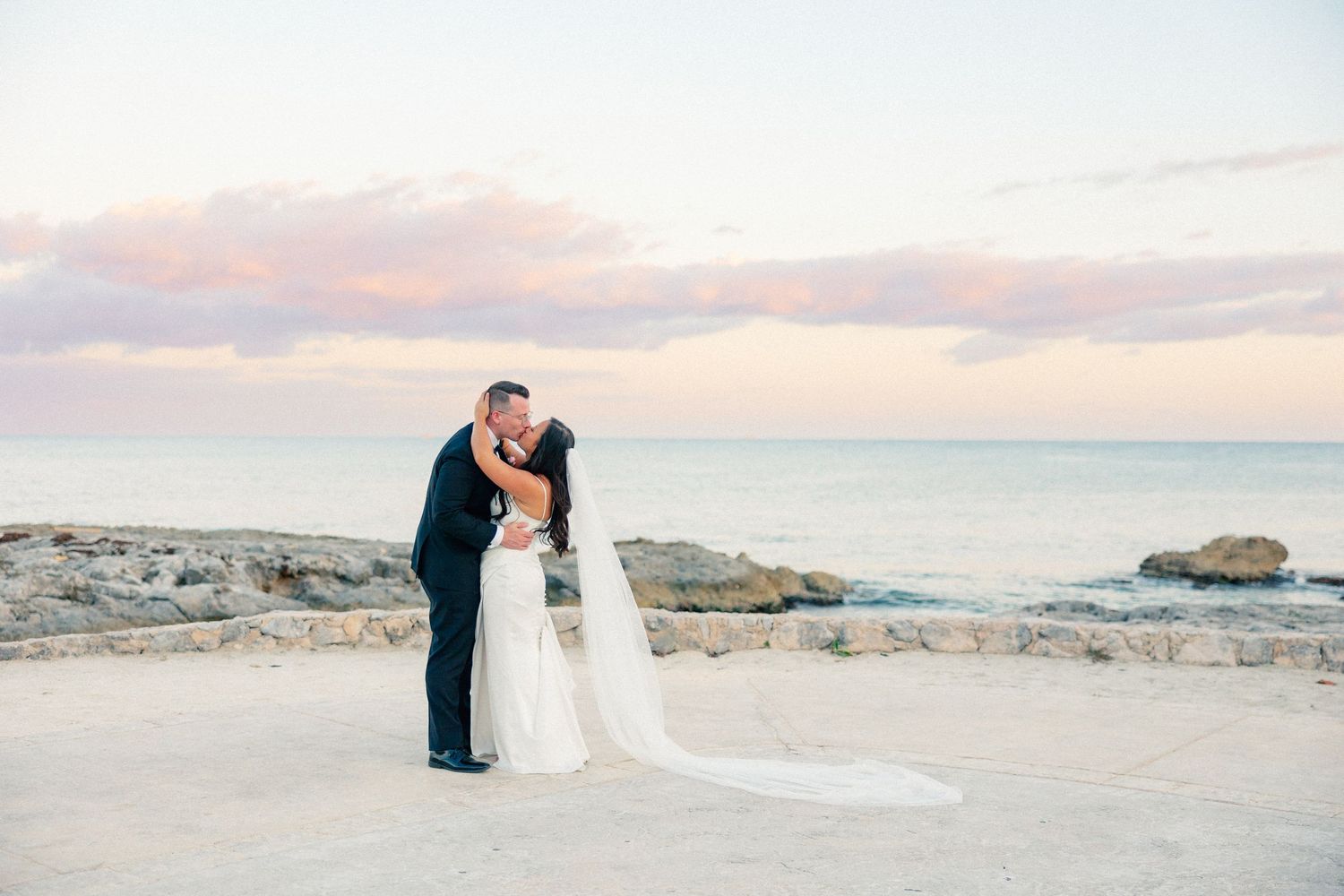 Bride and groom share a romantic kiss at sunset on a rocky oceanside terrace in a destination beach wedding.