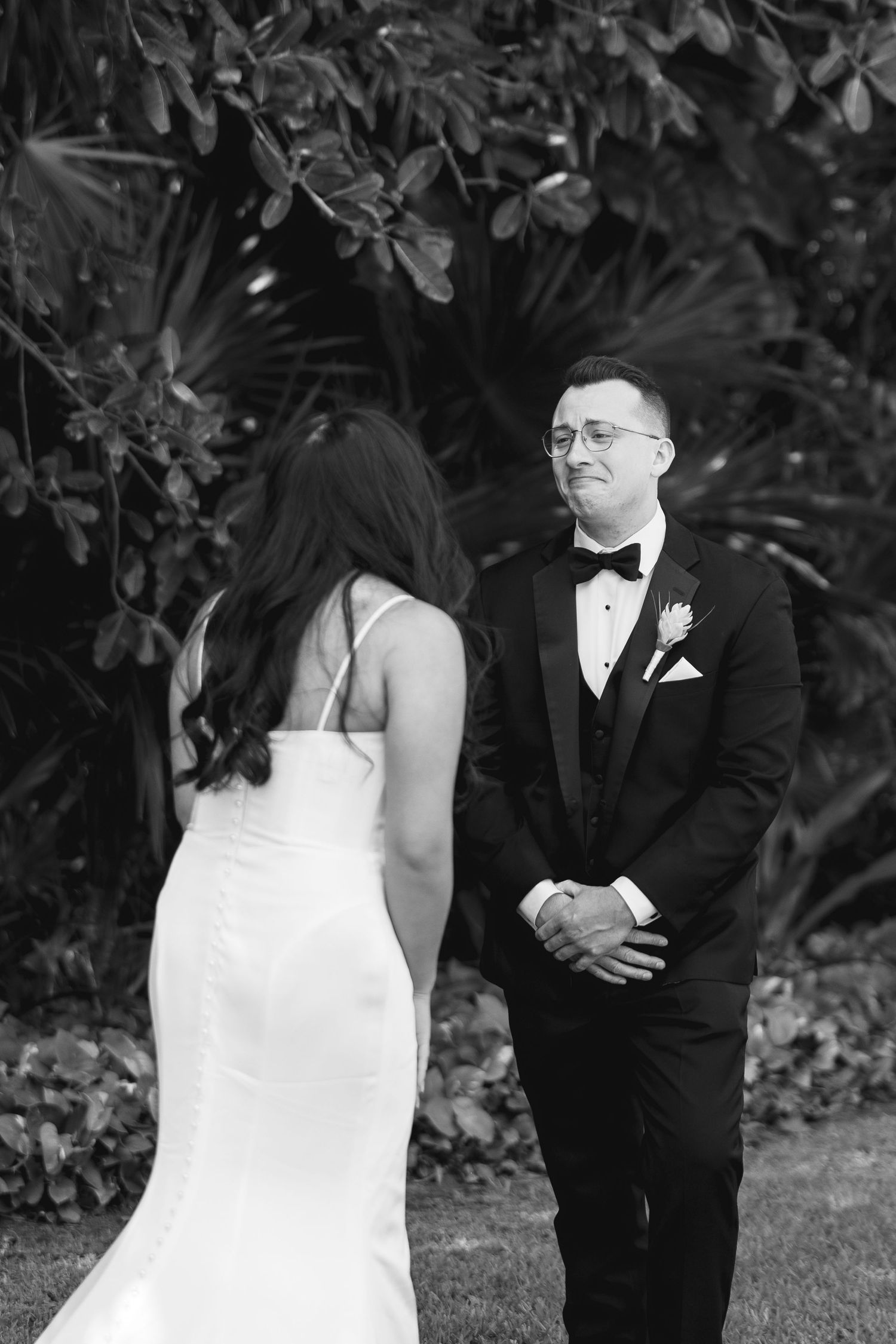 Black and white photo of groom seeing bride for first time at outdoor wedding ceremony with tropical foliage backdrop.