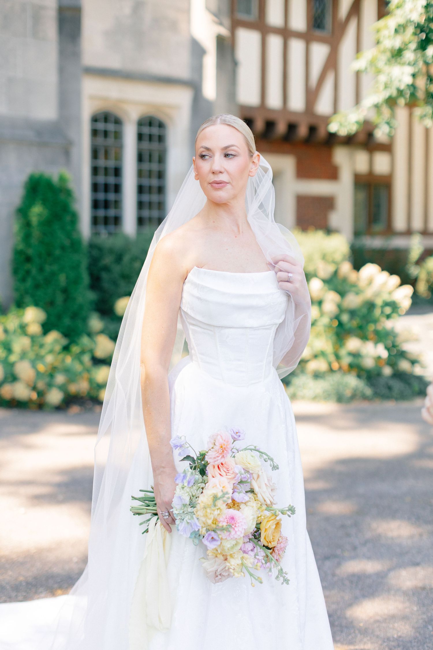 Bride in strapless white gown and veil holds pastel bouquet outdoors near Tudor-style building.