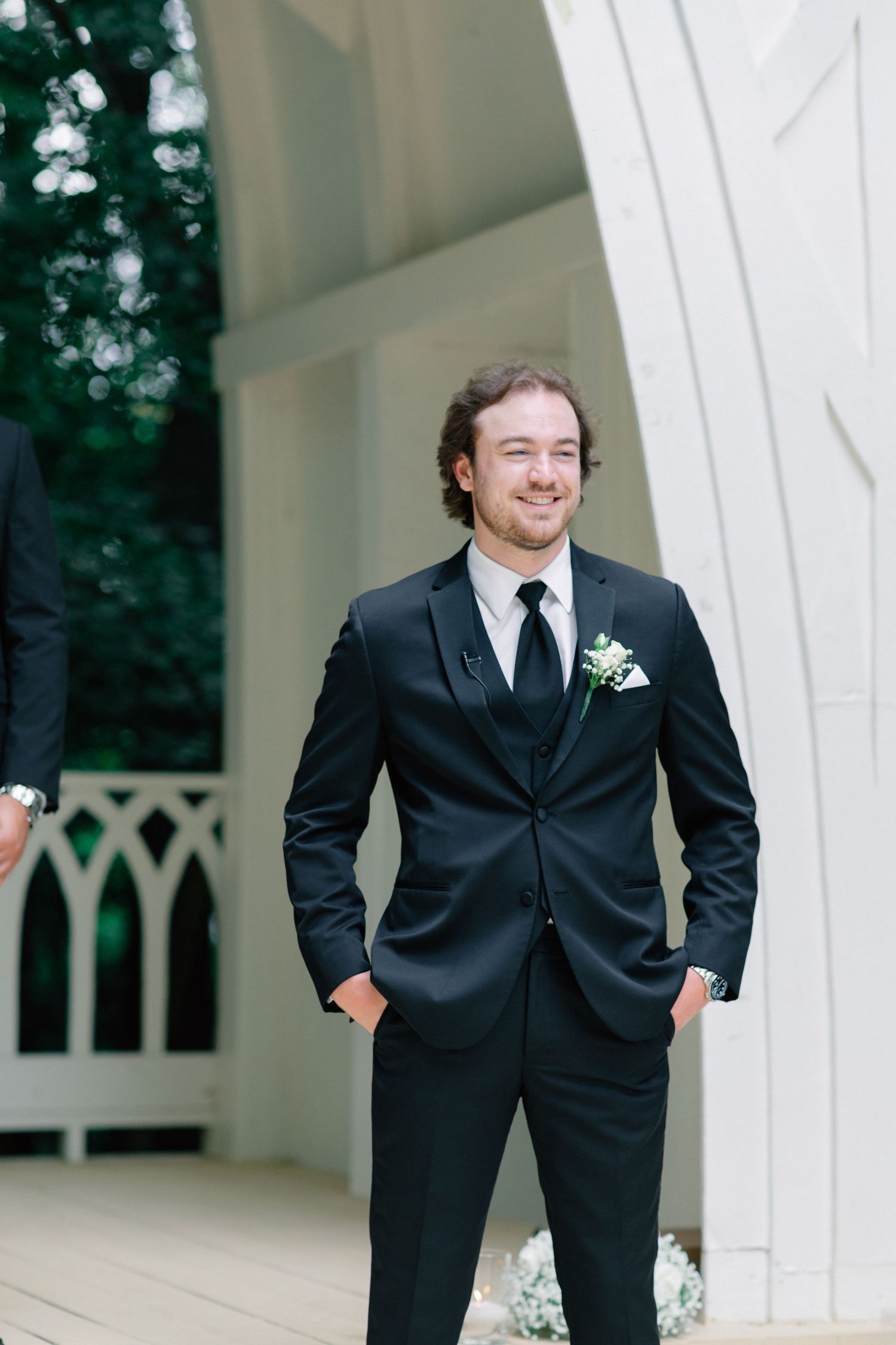 Smiling groom in black suit with white boutonniere stands confidently at outdoor wedding venue with white architectural details.