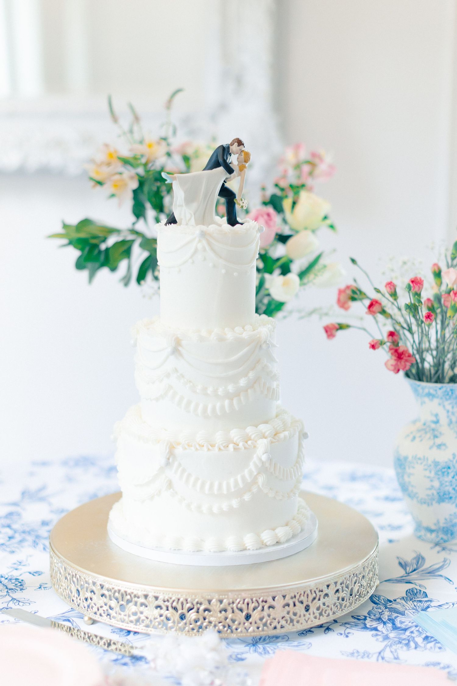 Elegant three-tier white wedding cake with bride and groom topper on silver stand, surrounded by pink and white floral arrangements, Florida