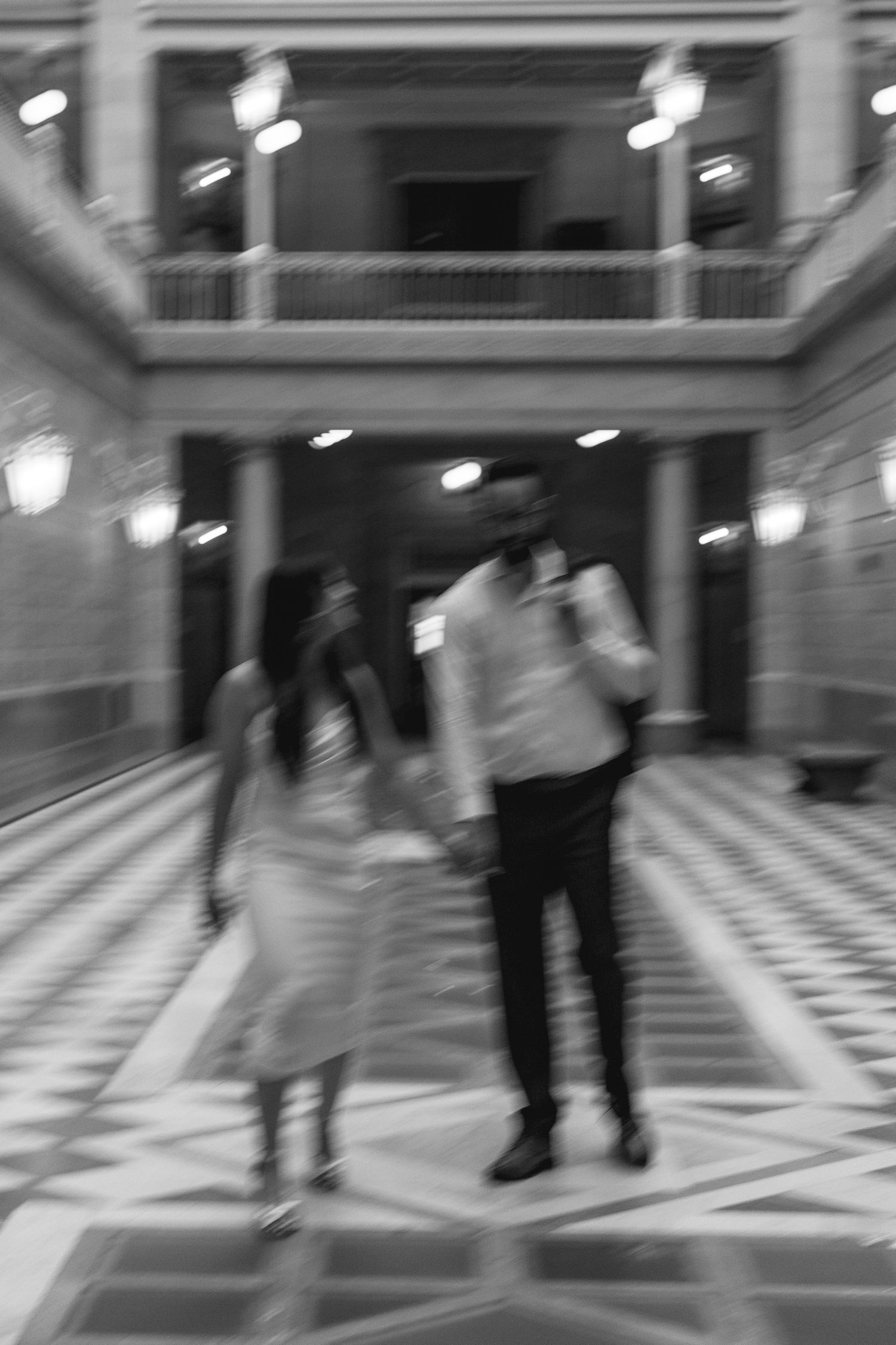 Blurred black and white photo of a couple walking hand in hand through a grand ornate building interior.