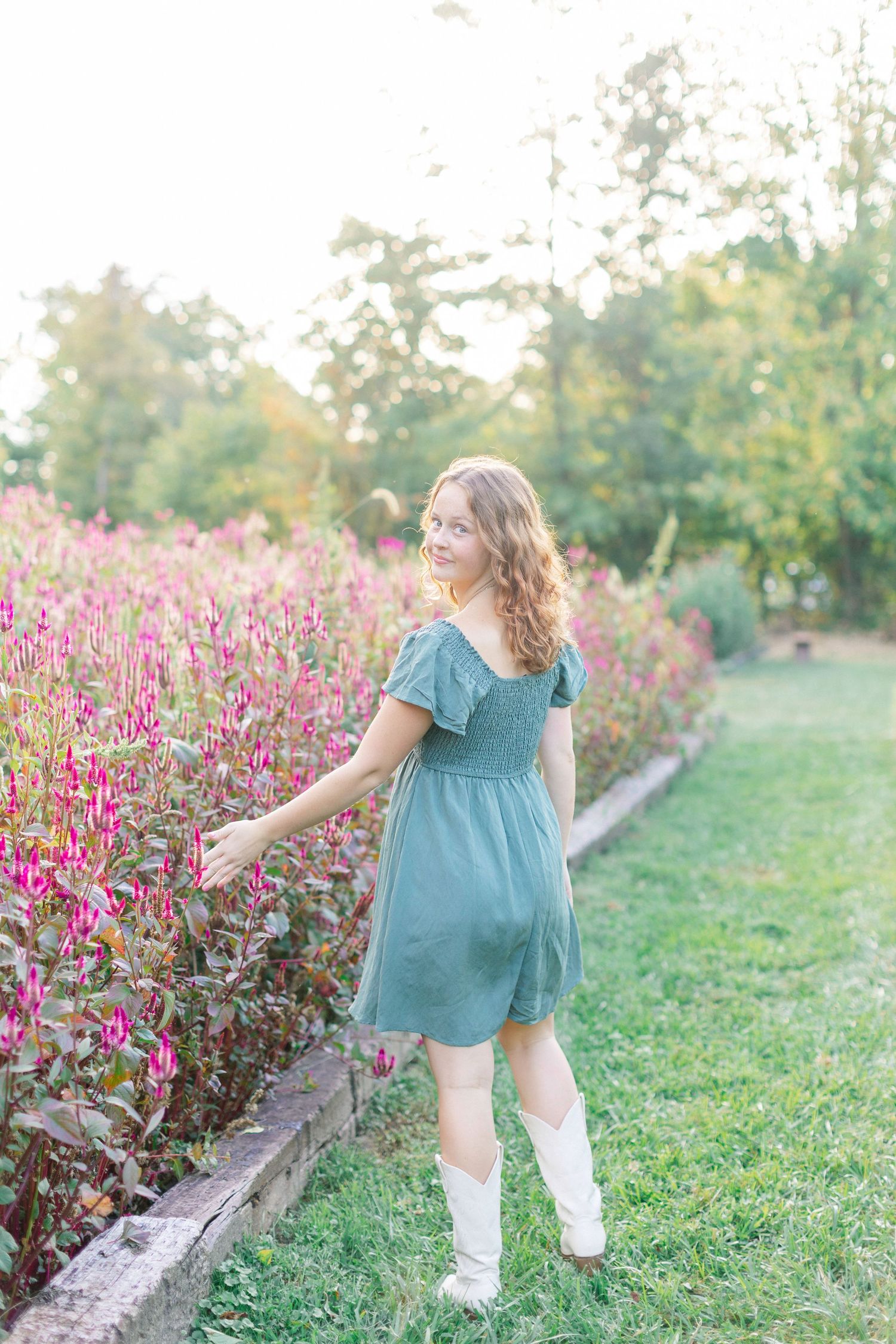 Young woman in teal dress and white boots smiling among vibrant pink flowers in a sunlit garden.
