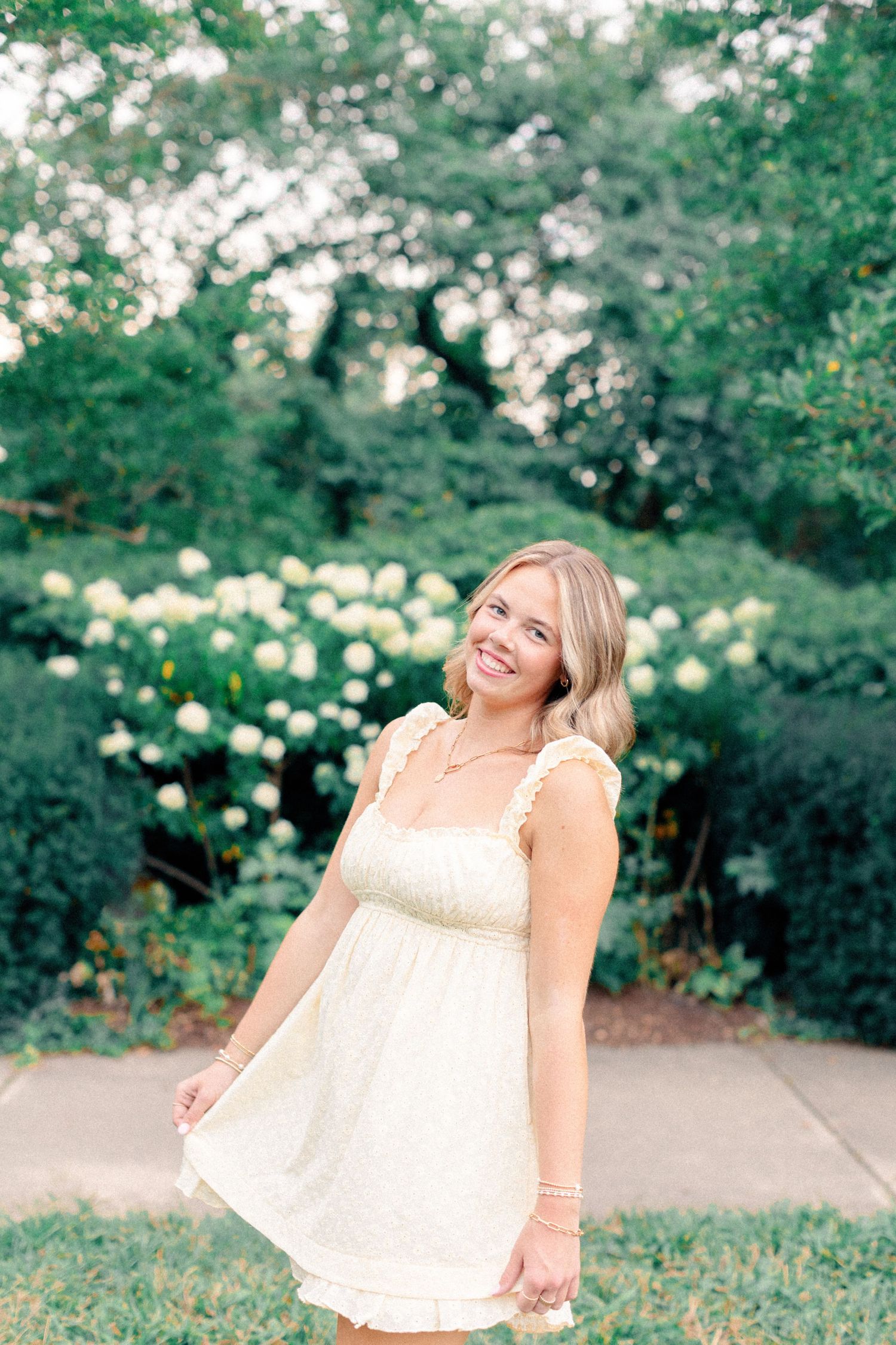 Cheerful blonde woman in a cream mini dress twirling in front of blooming hydrangea shrubs.