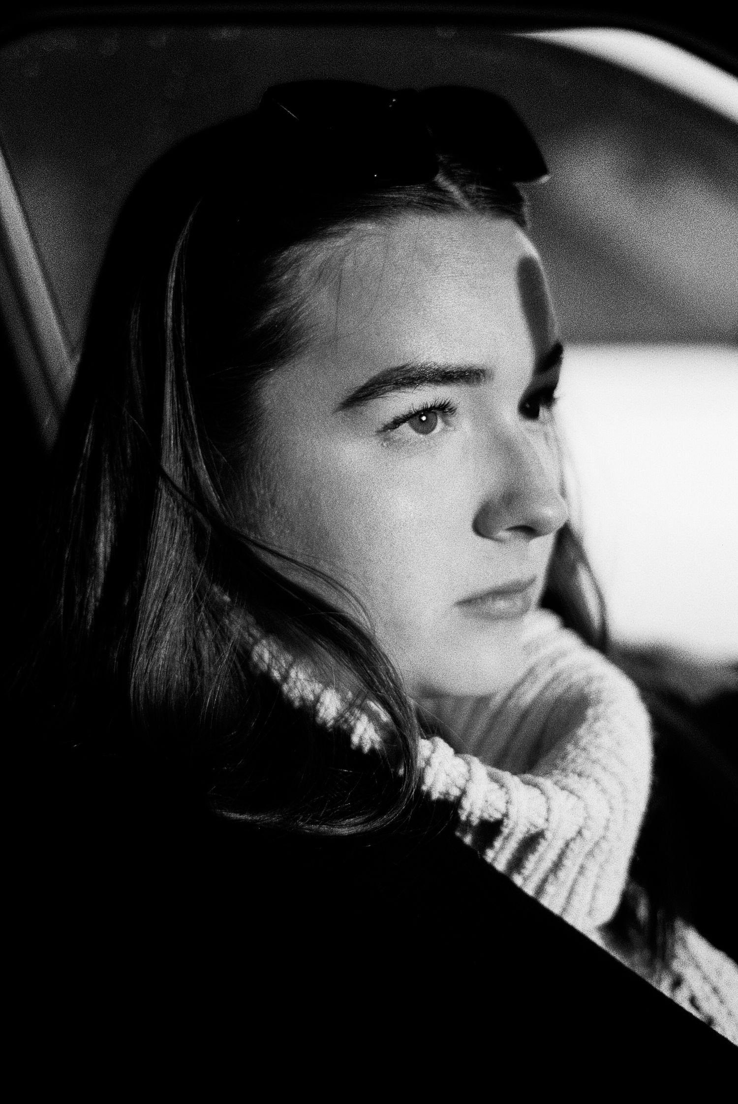 Black and white portrait of a young woman sitting in a car, wearing a knit sweater with sunglasses on her head.
