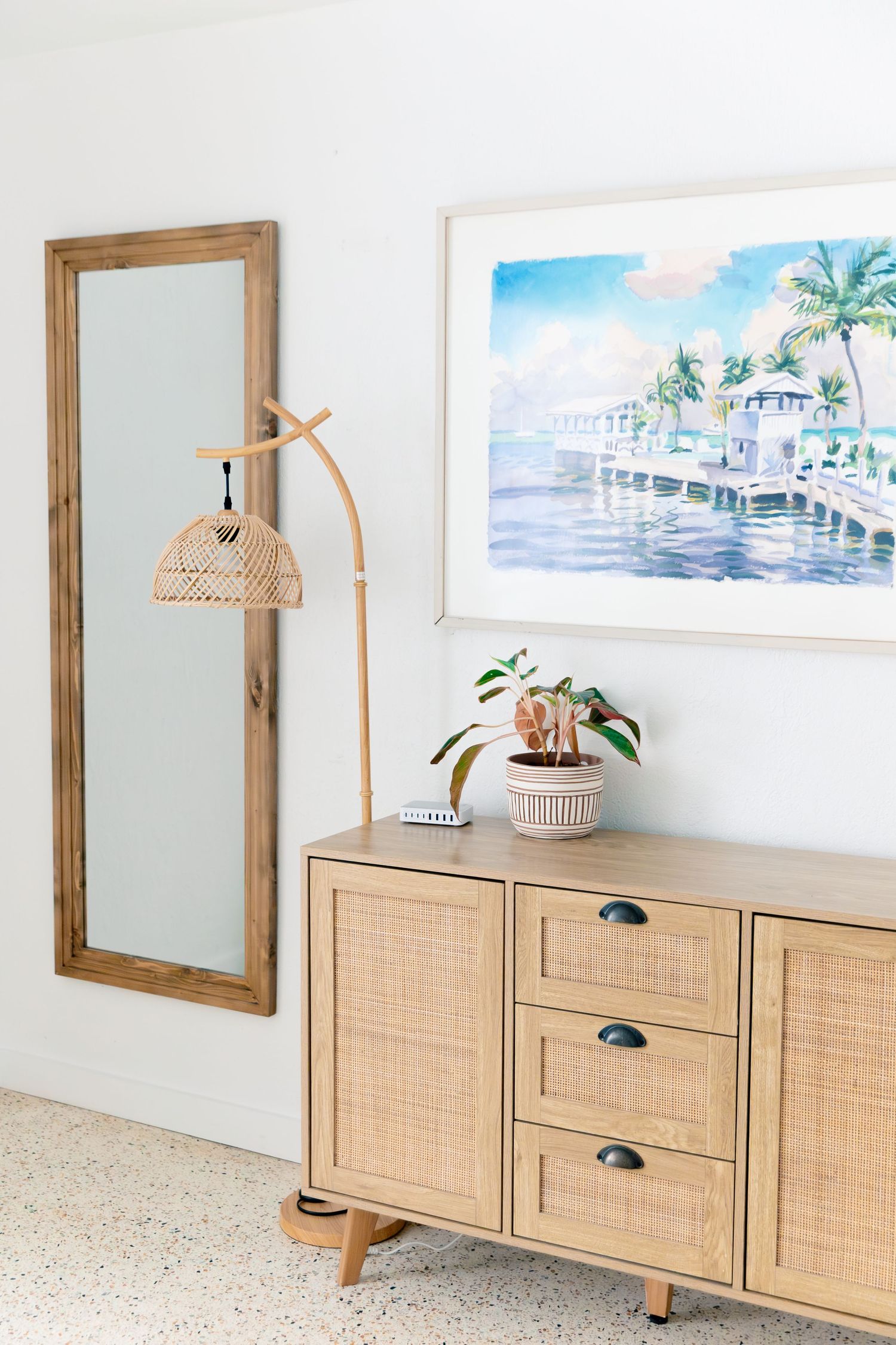 Coastal entryway with rattan sideboard, floor mirror, wicker lamp, and tropical watercolor artwork on white wall.
