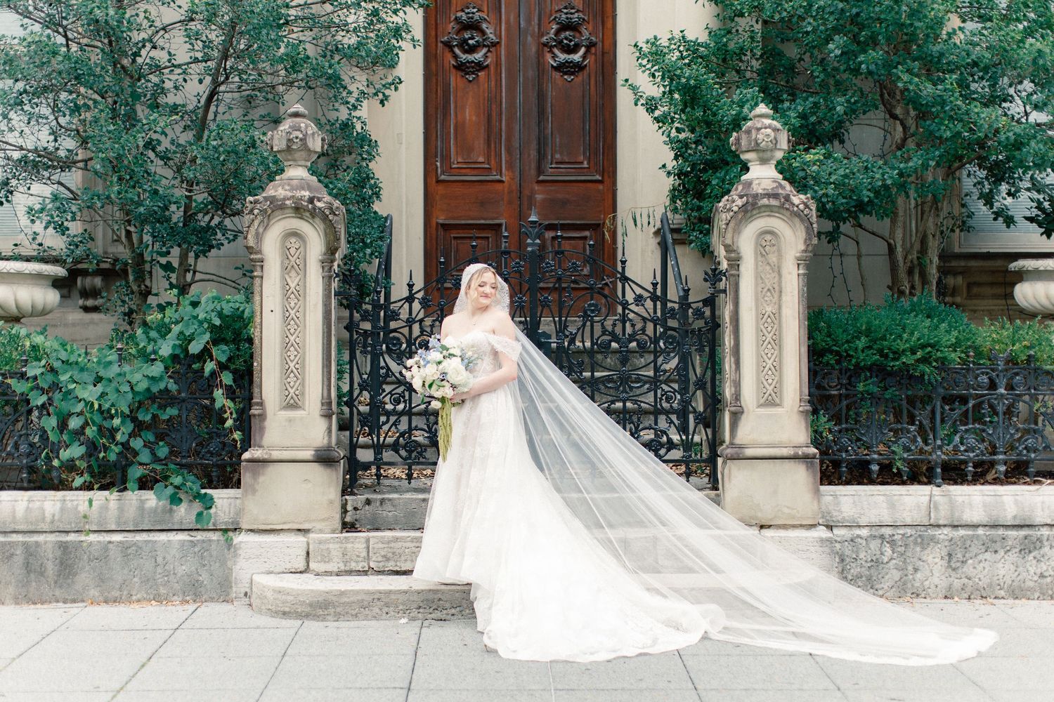 Bride in a flowing white gown and veil poses before ornate iron gates and a grand wooden door.