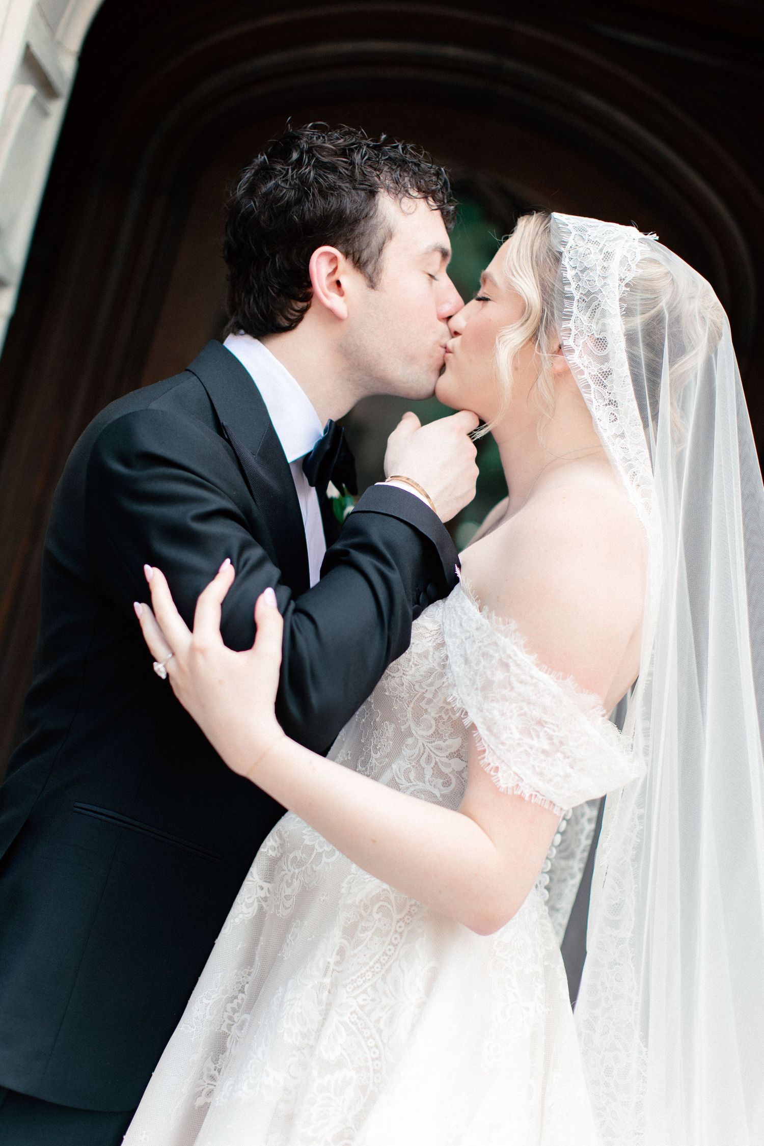 Bride and groom sharing a romantic kiss at church entrance, bride wearing lace wedding dress and veil.