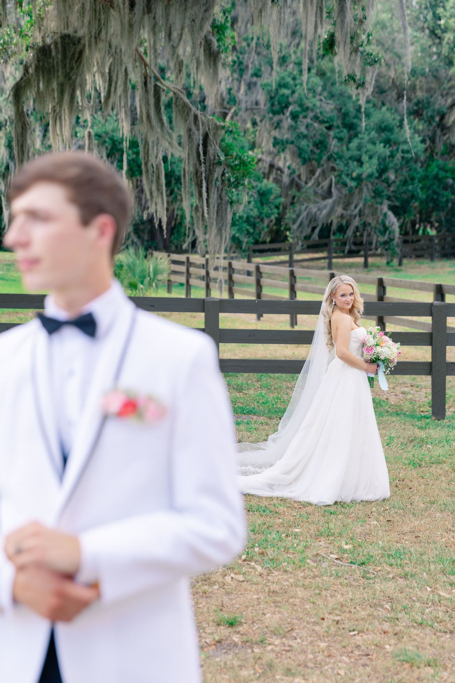 Bride in white gown holding bouquet approaches groom in white tuxedo during first look at rustic outdoor wedding venue, wedding Bella Cosa
