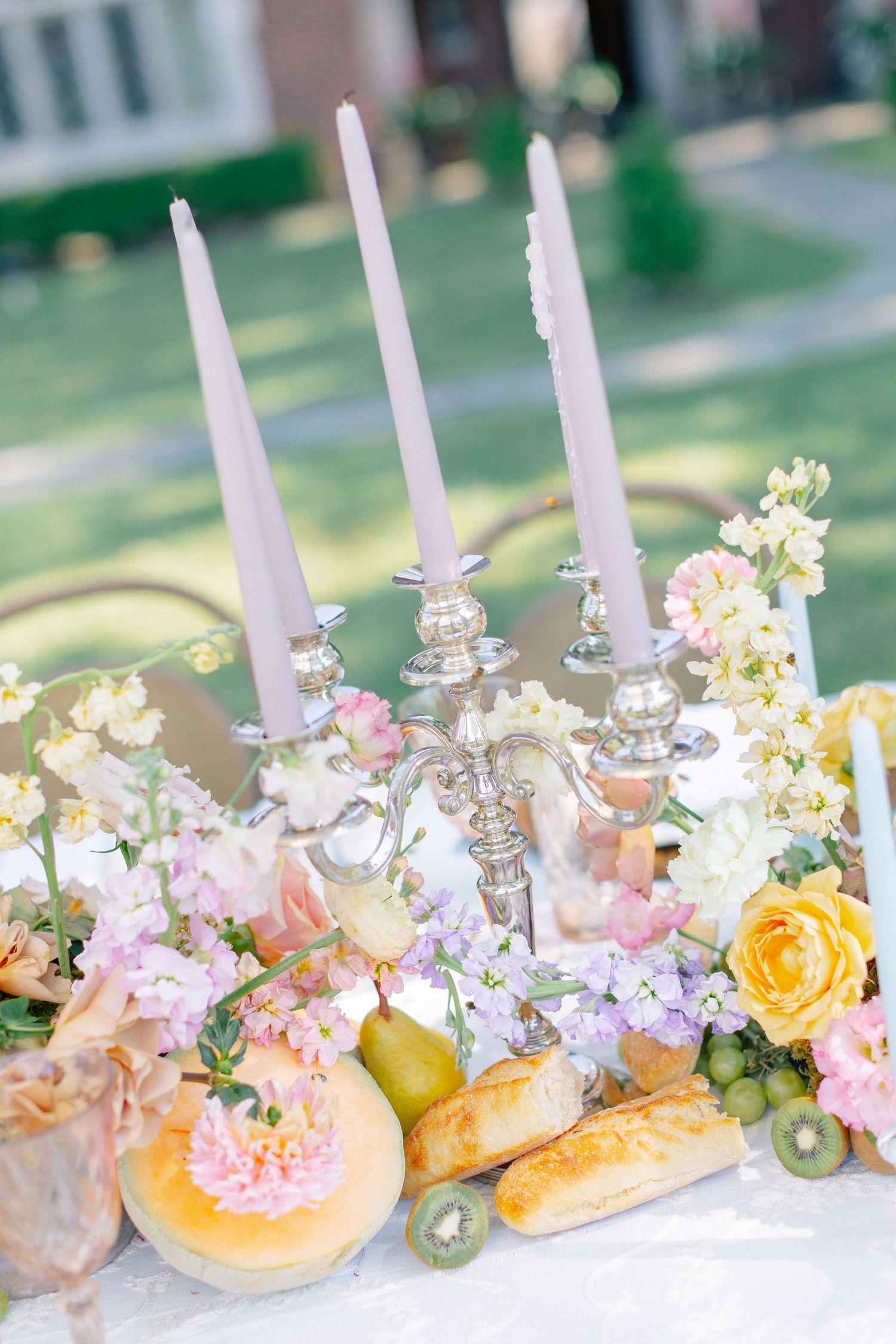 Outdoor wedding tablescape with silver candelabra, lavender taper candles, pastel flowers, and fresh fruit display.