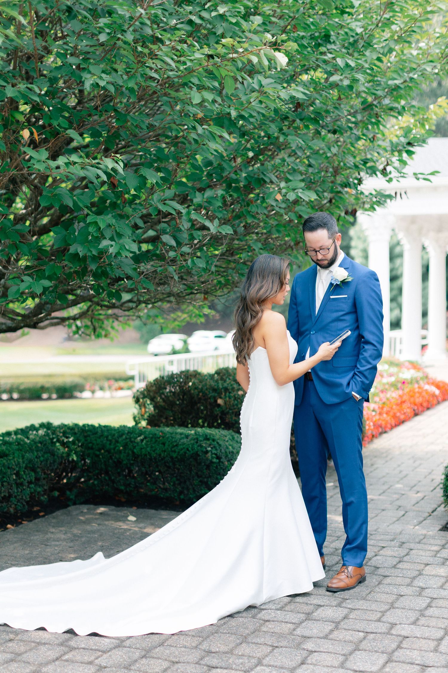 Bride in white gown and groom in blue suit share a tender moment on a garden patio with lush greenery.
