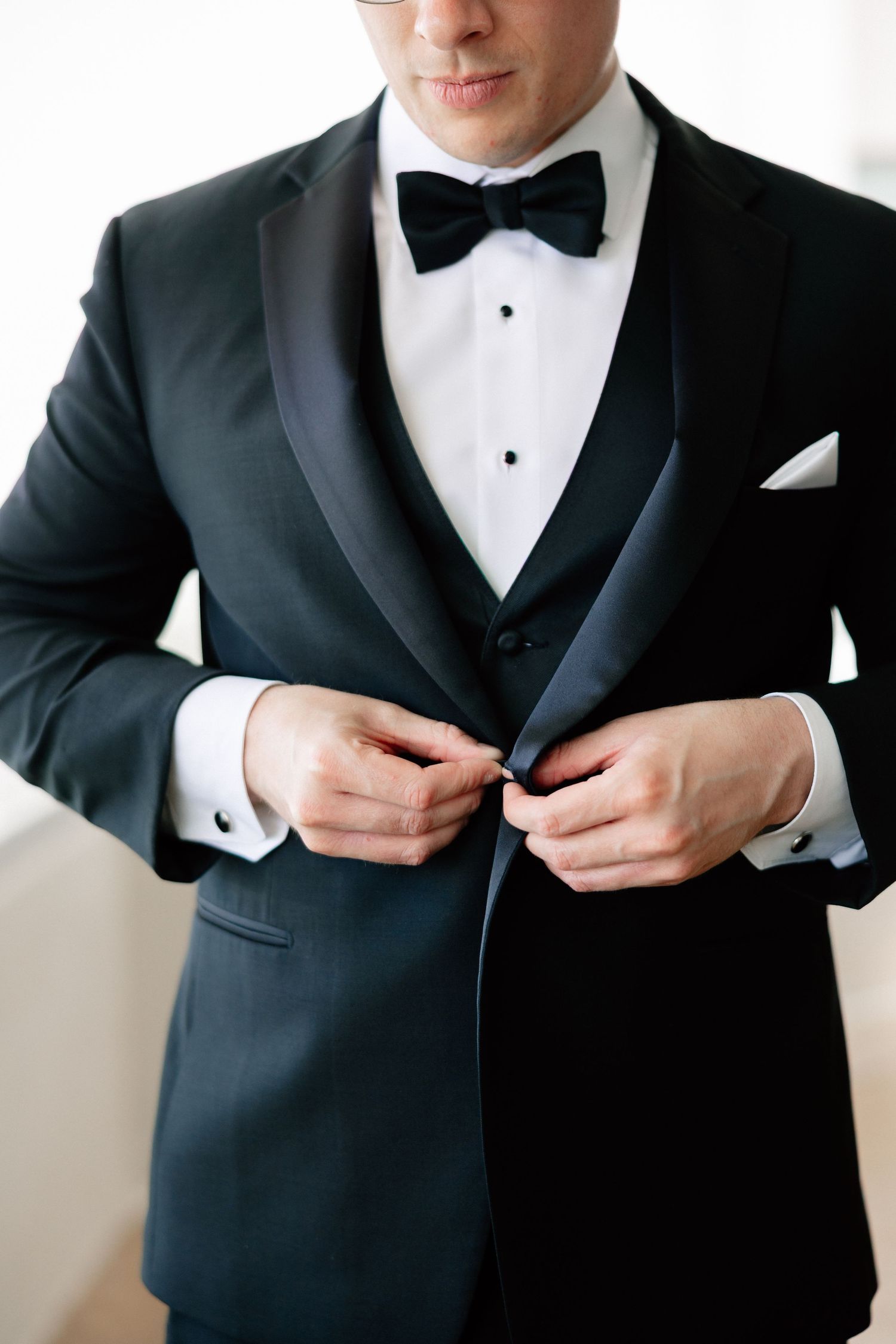 Man in black tuxedo with bow tie buttoning jacket, elegant formal attire for wedding or black tie event.