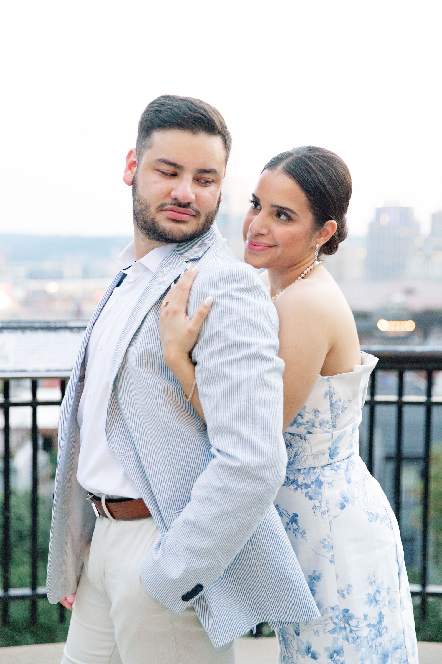 Bride in a blue floral strapless gown embraces groom from behind as they pose together on a rooftop overlook.