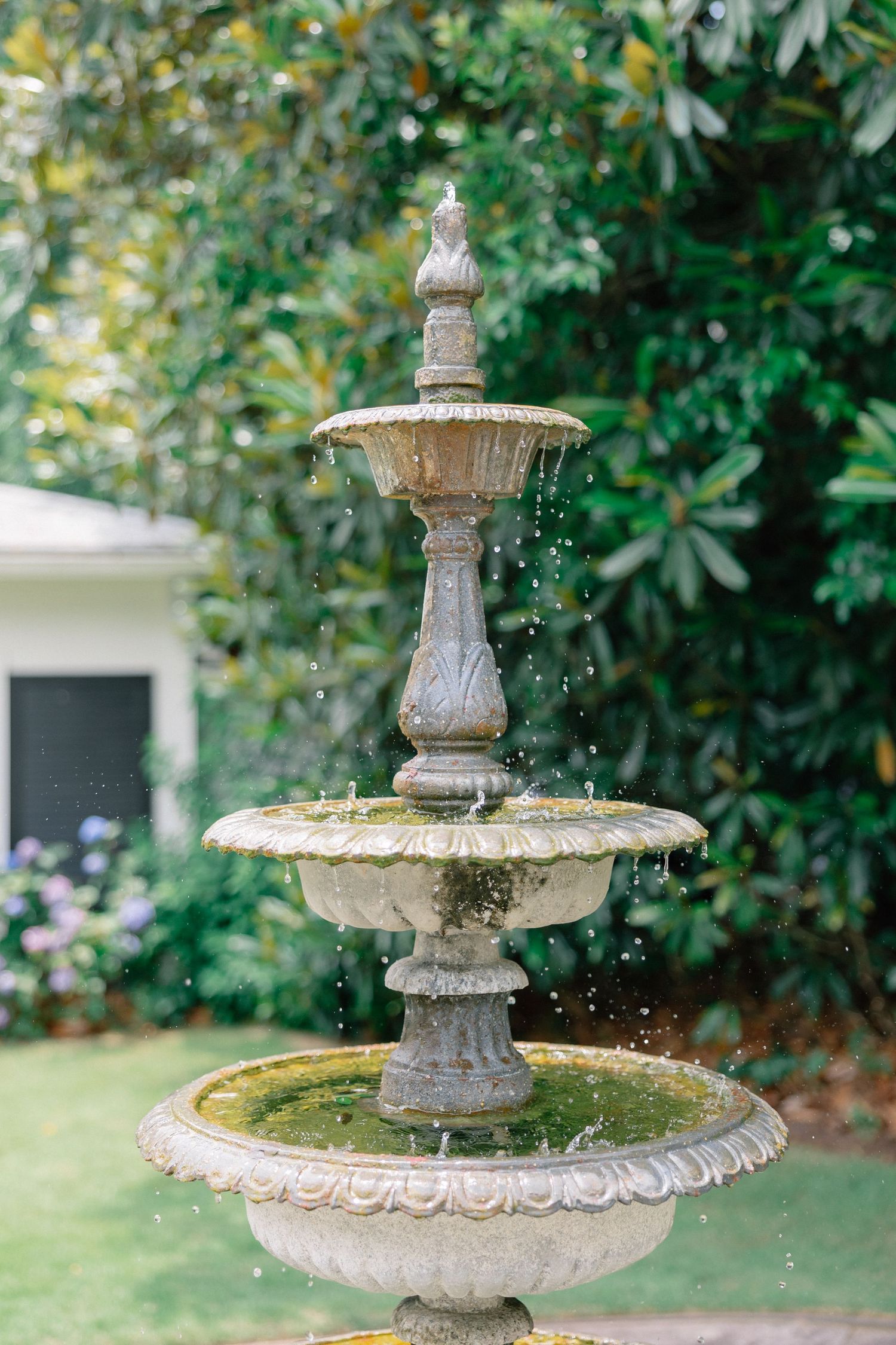 A classic three-tiered stone garden fountain with cascading water, surrounded by lush green trees and purple flowers.