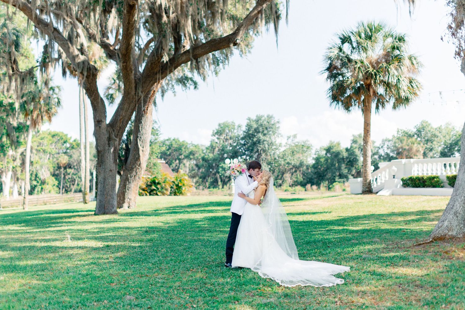 Bride and groom share a romantic kiss on lush green lawn surrounded by Spanish moss-draped oak and palm trees, wedding at Bella Cosa