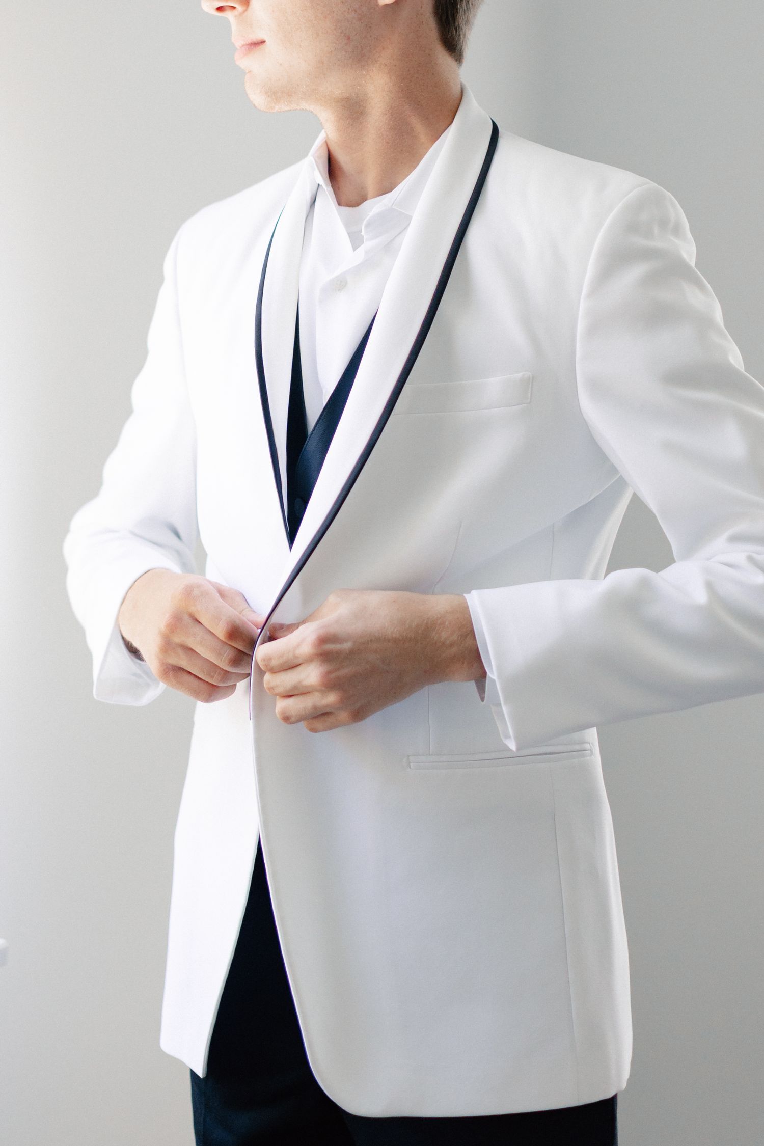 Man buttoning a white tuxedo jacket with black lapel trim, styled with a dark tie and white dress shirt, wedding at Bella Cosa