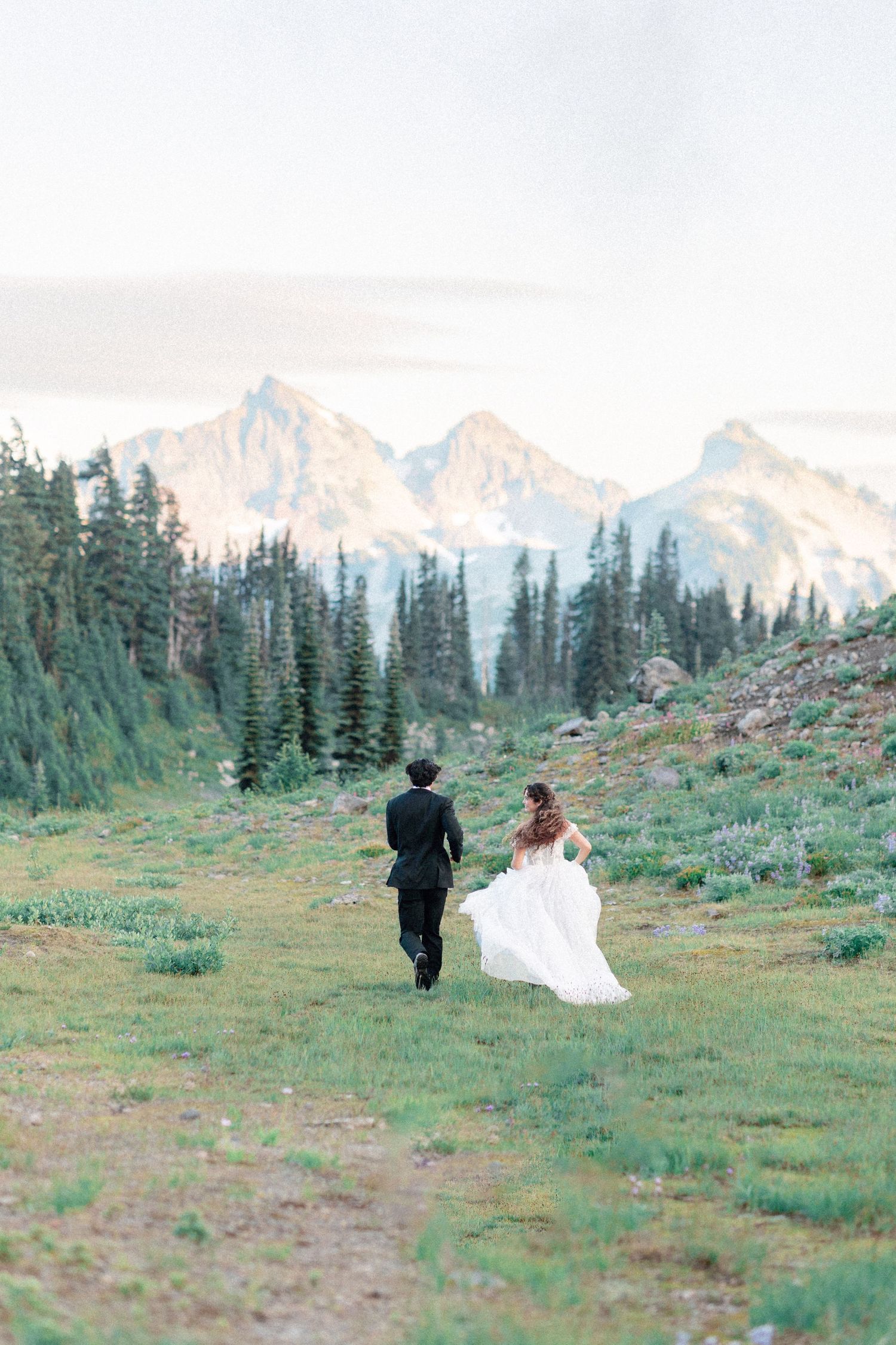 Bride and groom walking through alpine meadow with snow-capped mountains and evergreen trees in background.