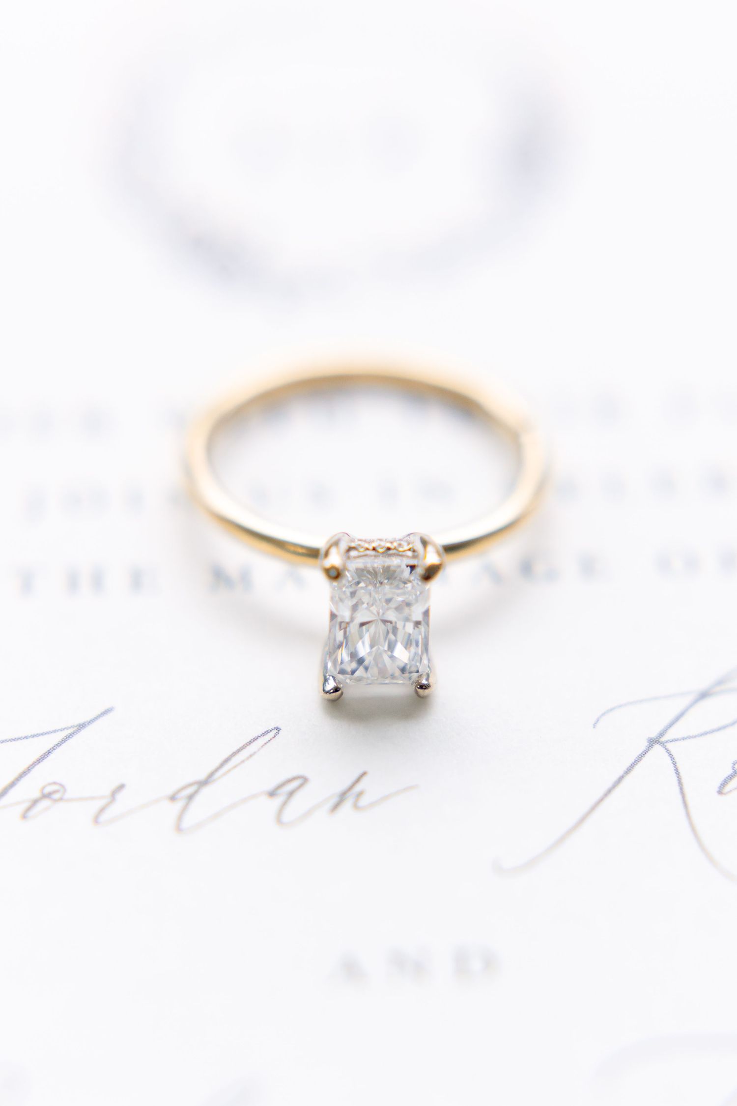 Gold solitaire engagement ring with emerald-cut diamond displayed on elegant wedding invitation stationery.
