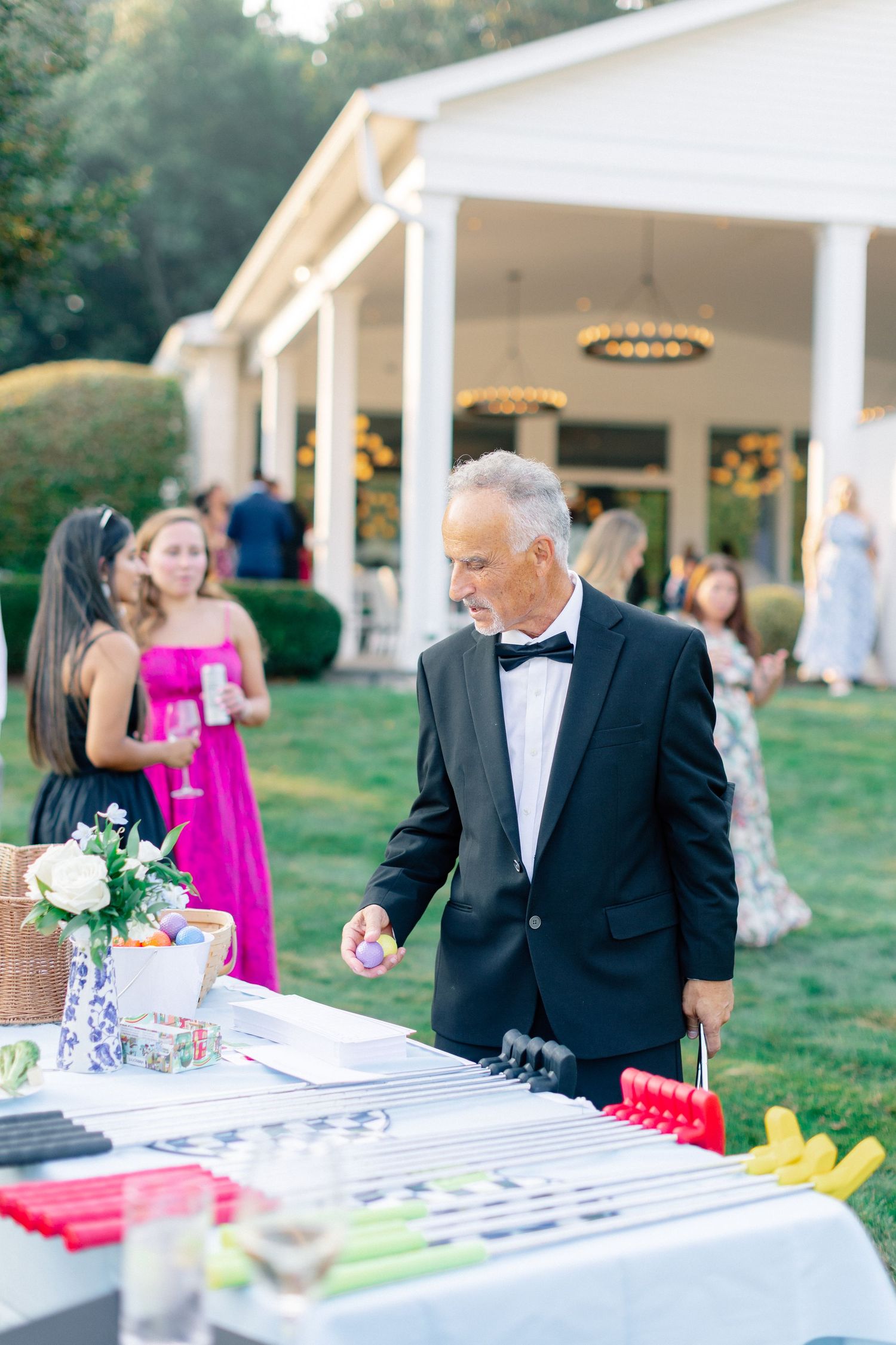 Elegant man in tuxedo at outdoor wedding cocktail hour, standing at decorated table with party favors on manicured lawn.