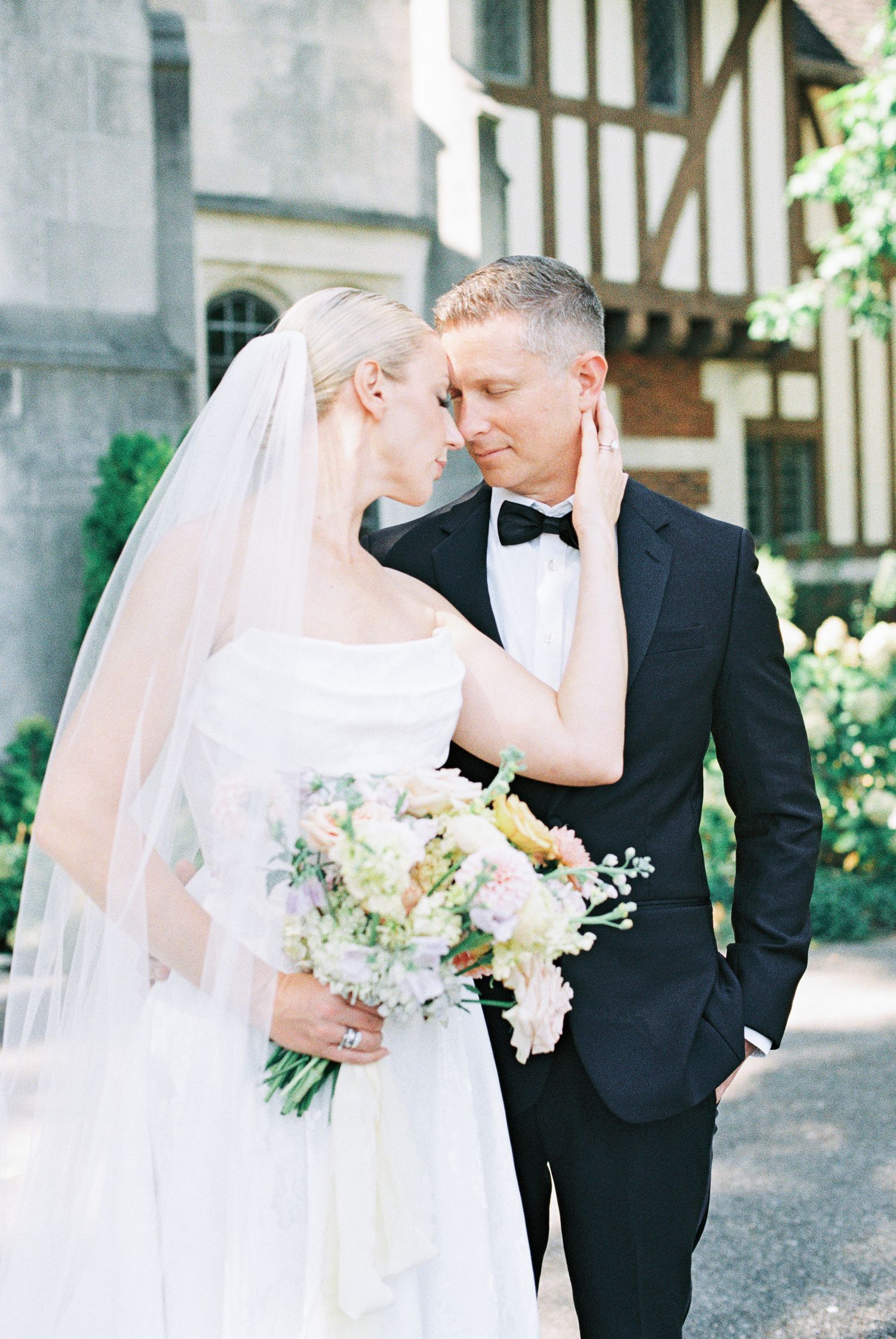 Bride and groom share an intimate moment, touching foreheads outside a historic Tudor-style building.