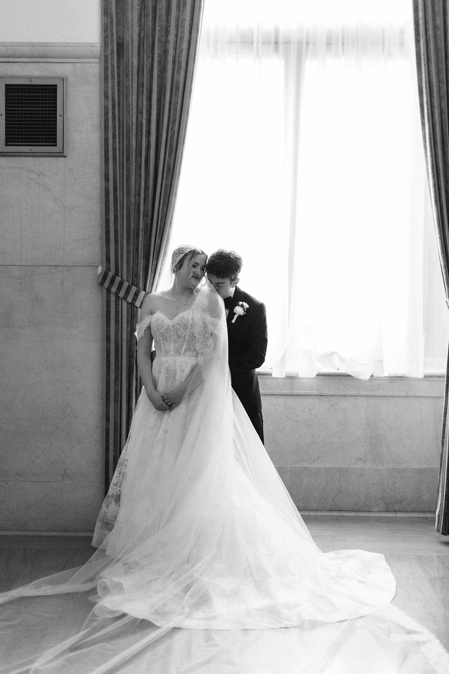 Black and white wedding photo of bride in flowing gown and groom in tuxedo standing by draped window.