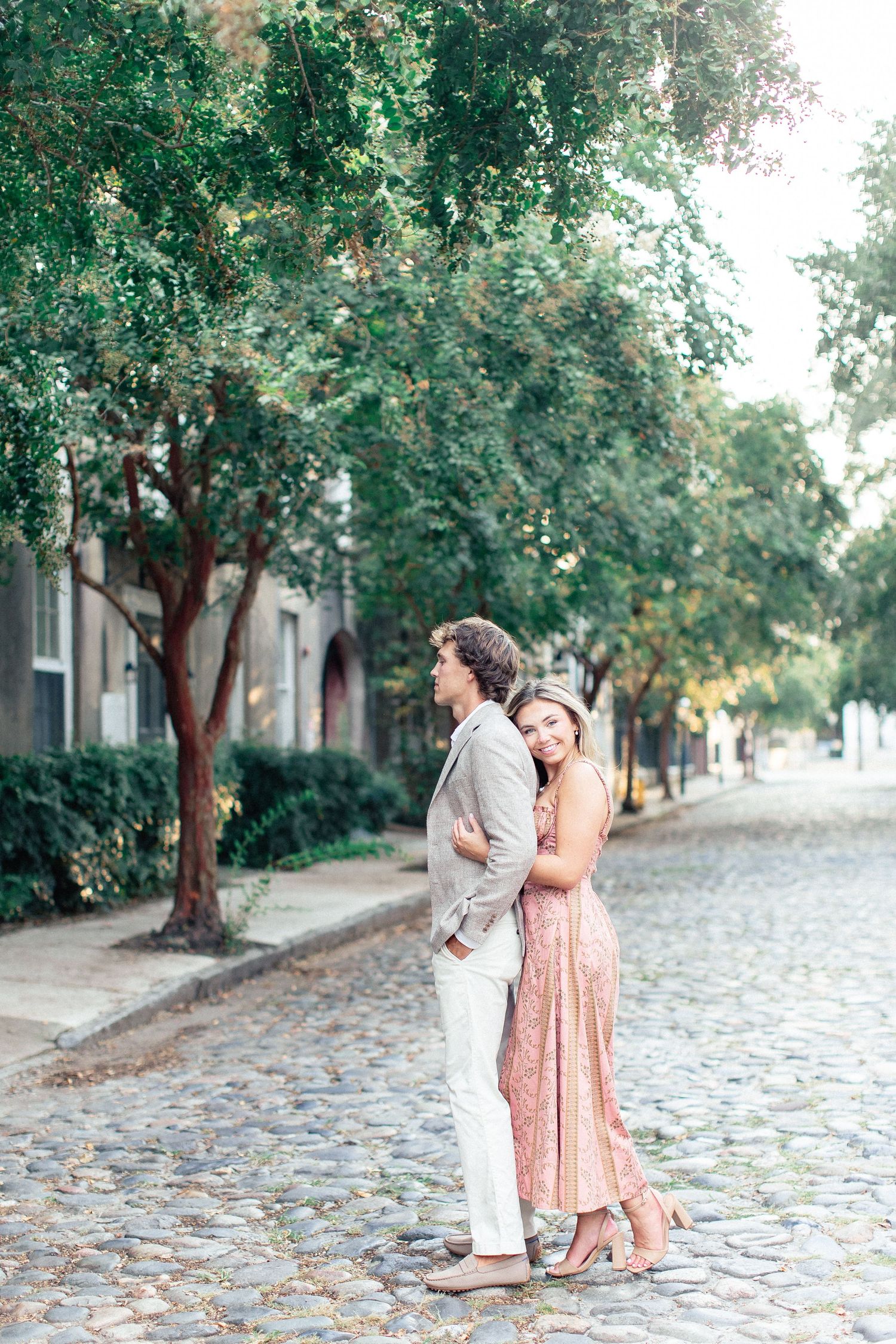 Couple embracing on a charming cobblestone street lined with green trees, wearing elegant casual attire.