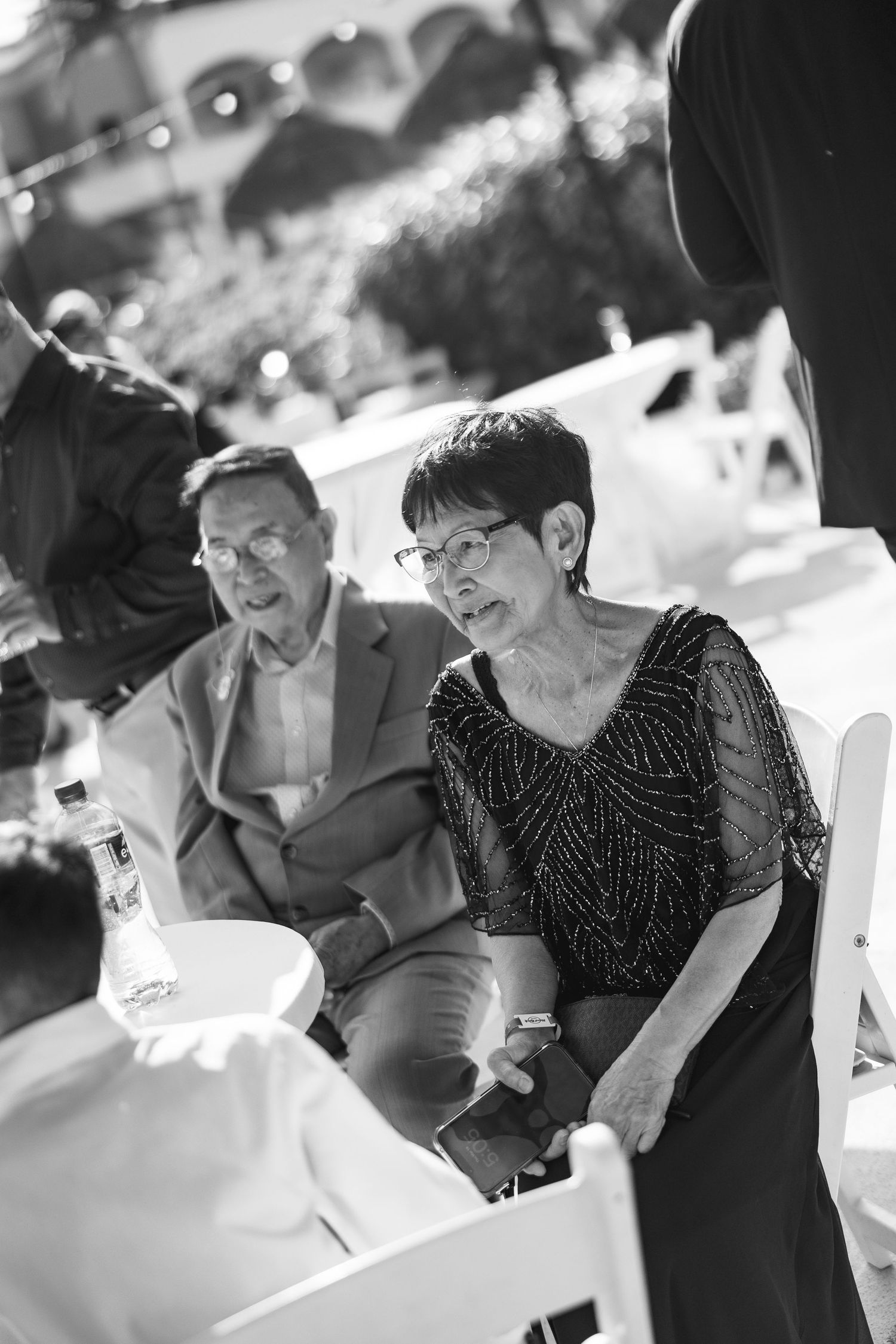 Black and white photo of elderly couple seated at outdoor wedding reception, woman in beaded dress smiling.