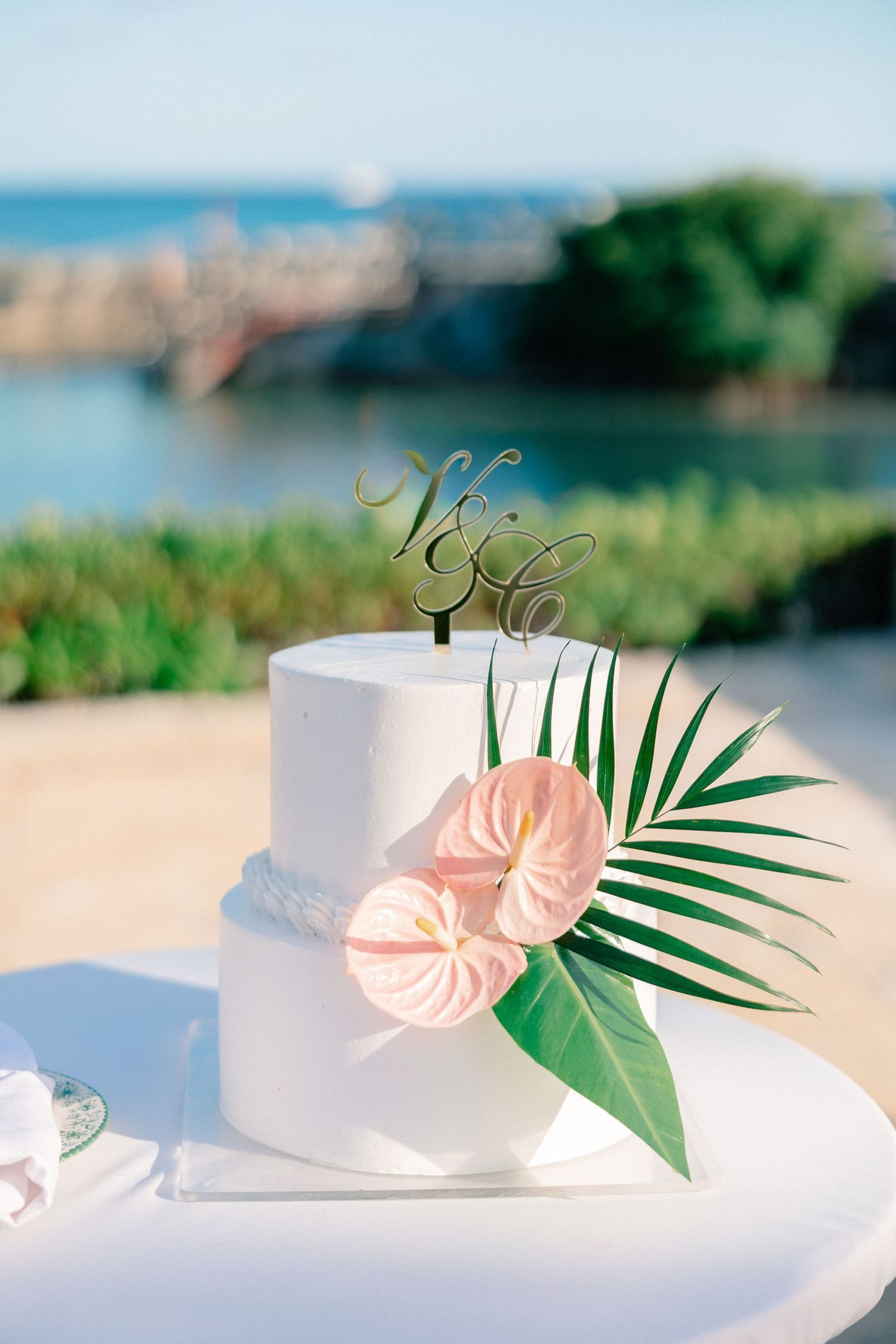 Elegant two-tier white wedding cake with pink anthurium, tropical palm leaf, and gold monogram topper by ocean.
