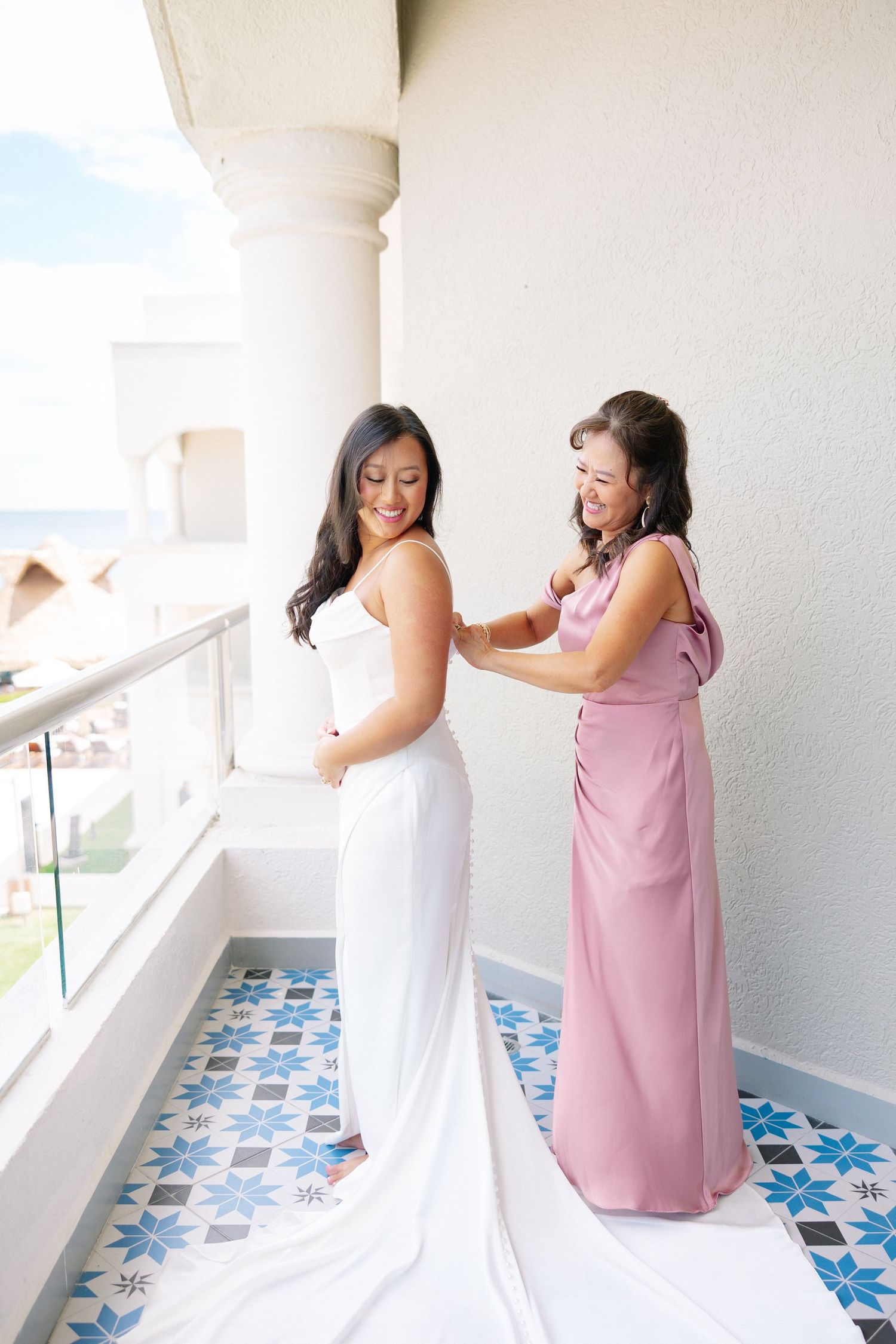 Bride in white gown gets dress fastened by bridesmaid in pink on a balcony with decorative tile flooring.
