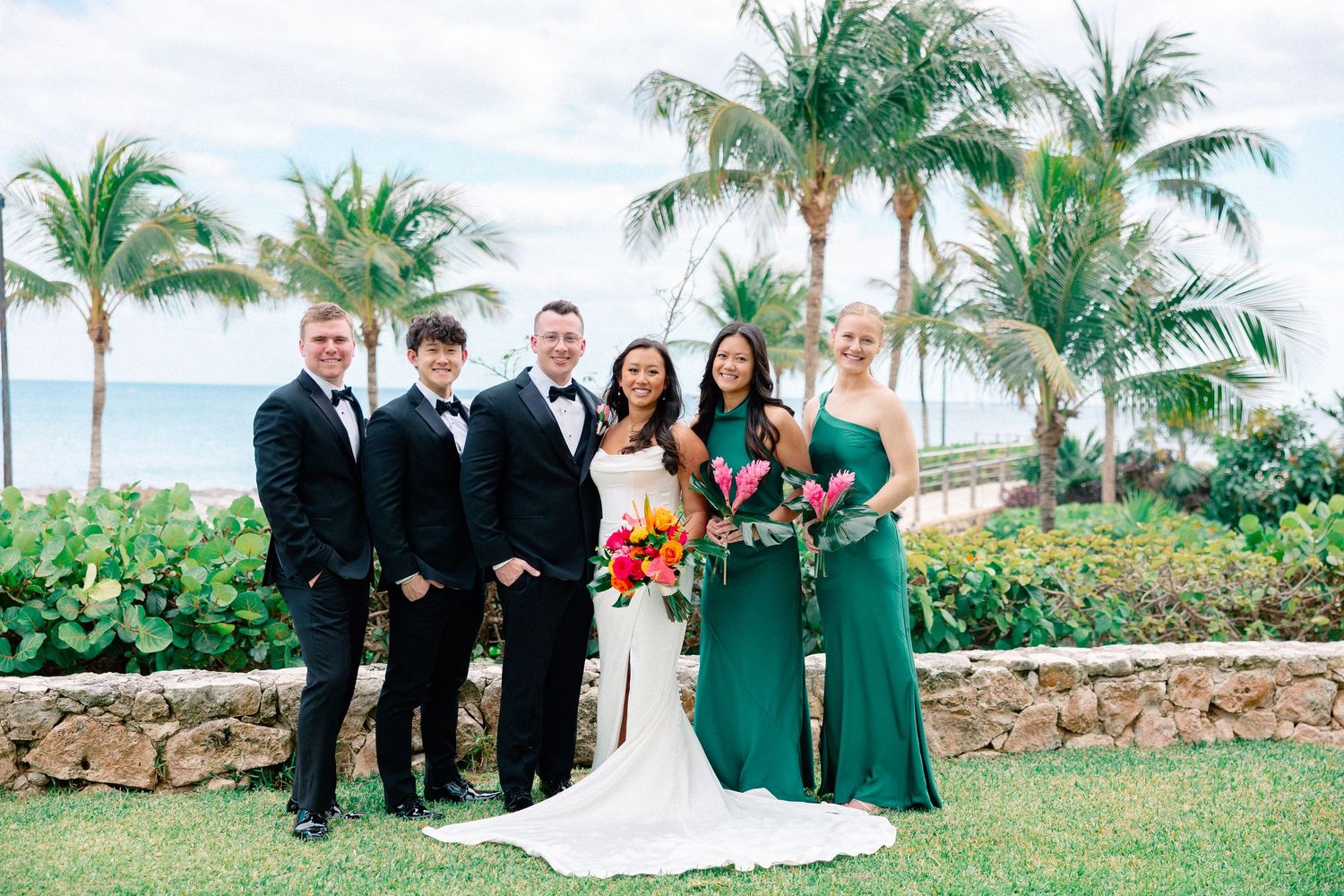 Wedding party posing outdoors near palm trees and ocean, bride in white gown with colorful bouquet, groomsmen in tuxedos.