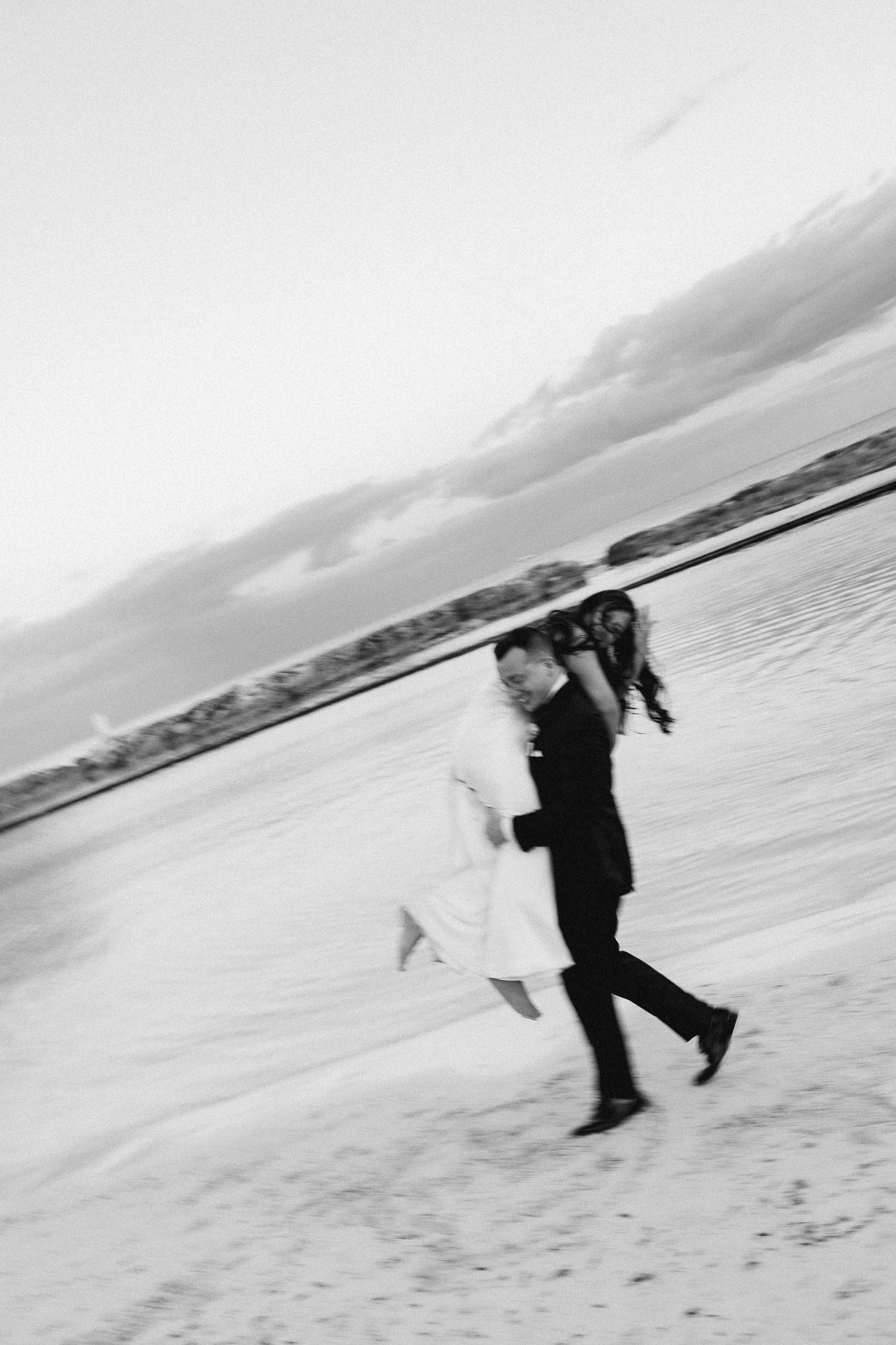 Black and white photo of a bride and groom running joyfully on a sandy beach, motion blur capturing their wedding day excitement.