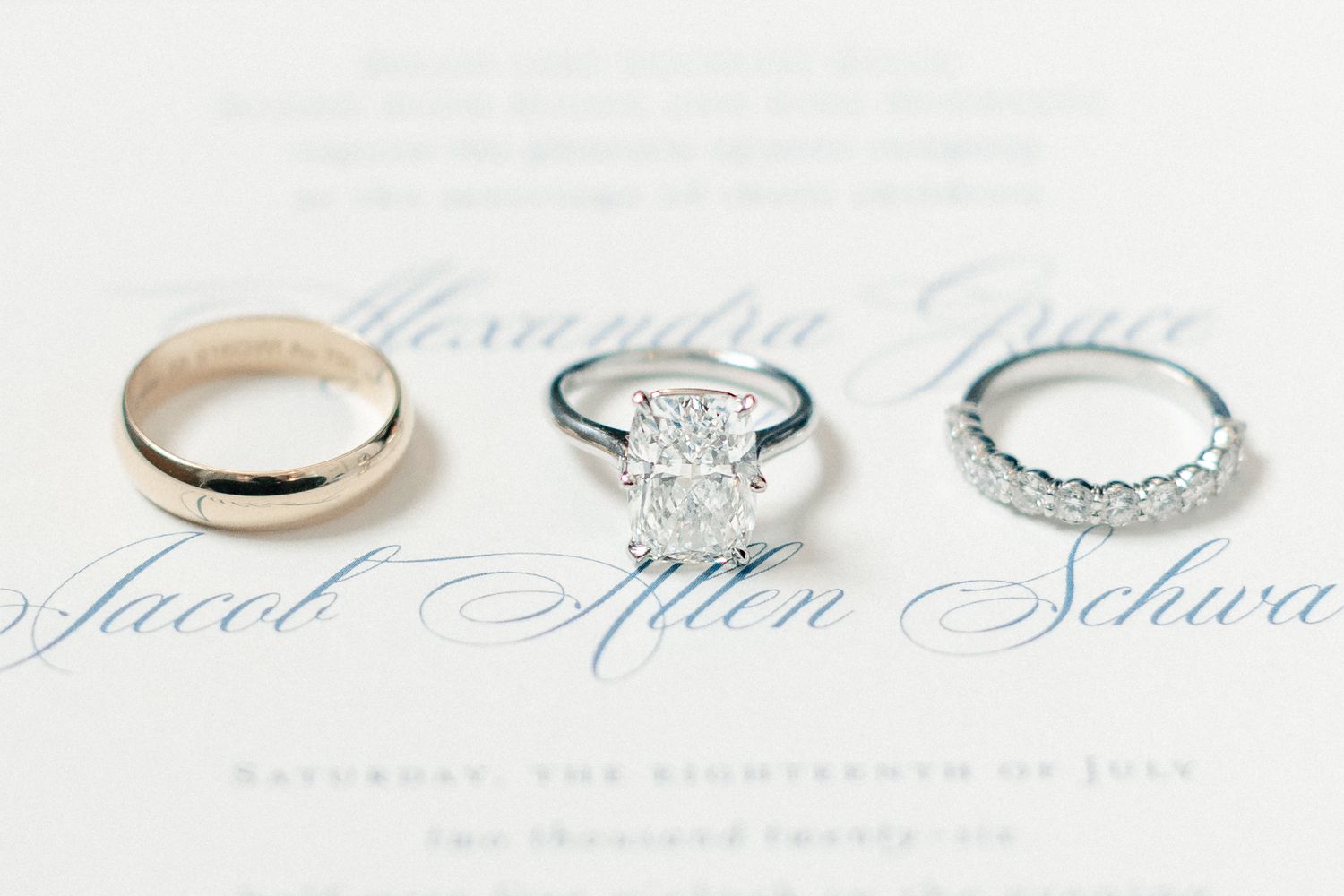 Three wedding rings displayed on elegant invitation, featuring gold band, oval diamond solitaire, and diamond eternity band.