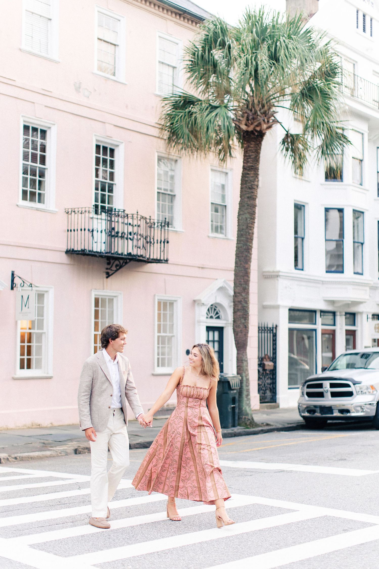 Engaged couple crossing a zebra crosswalk hand in hand near a pink building and palm tree in Charleston, SC.