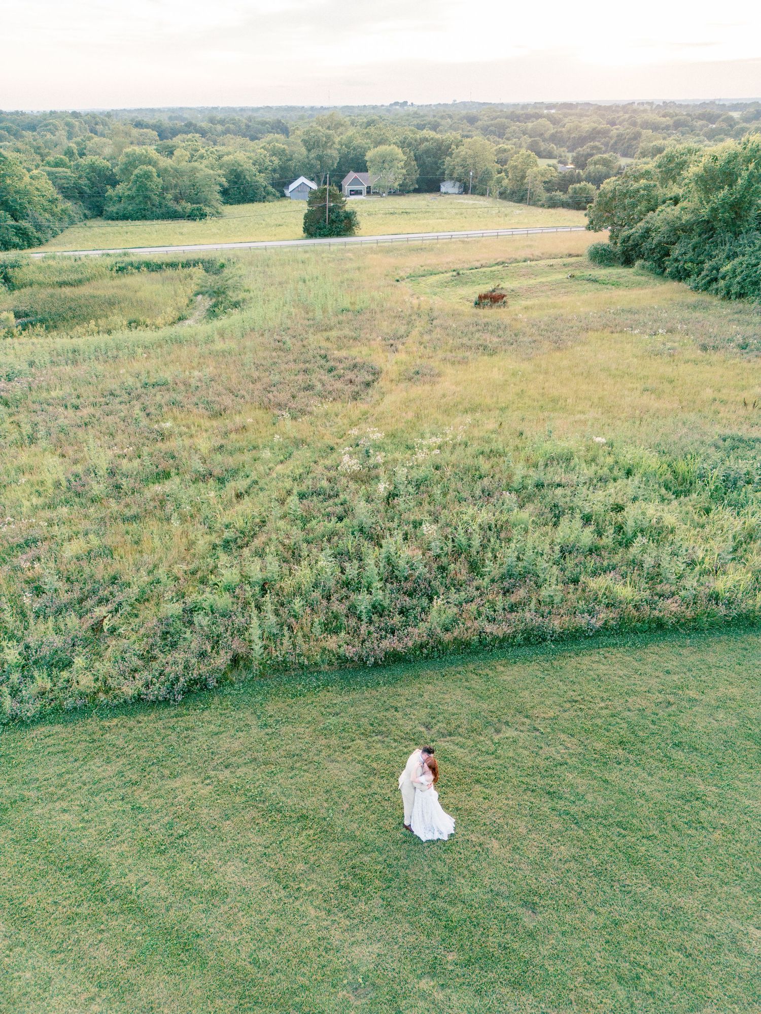 Aerial drone view of a bride and groom standing in a lush green field at golden hour on their wedding day.