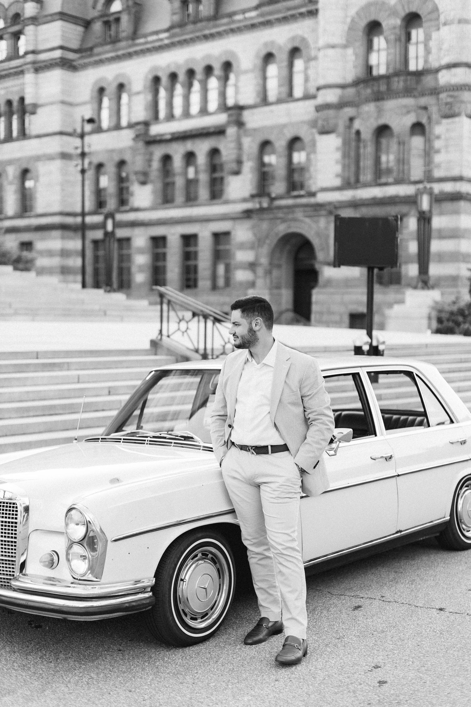 Man in light suit leaning against classic vintage Mercedes-Benz car in front of historic stone building.