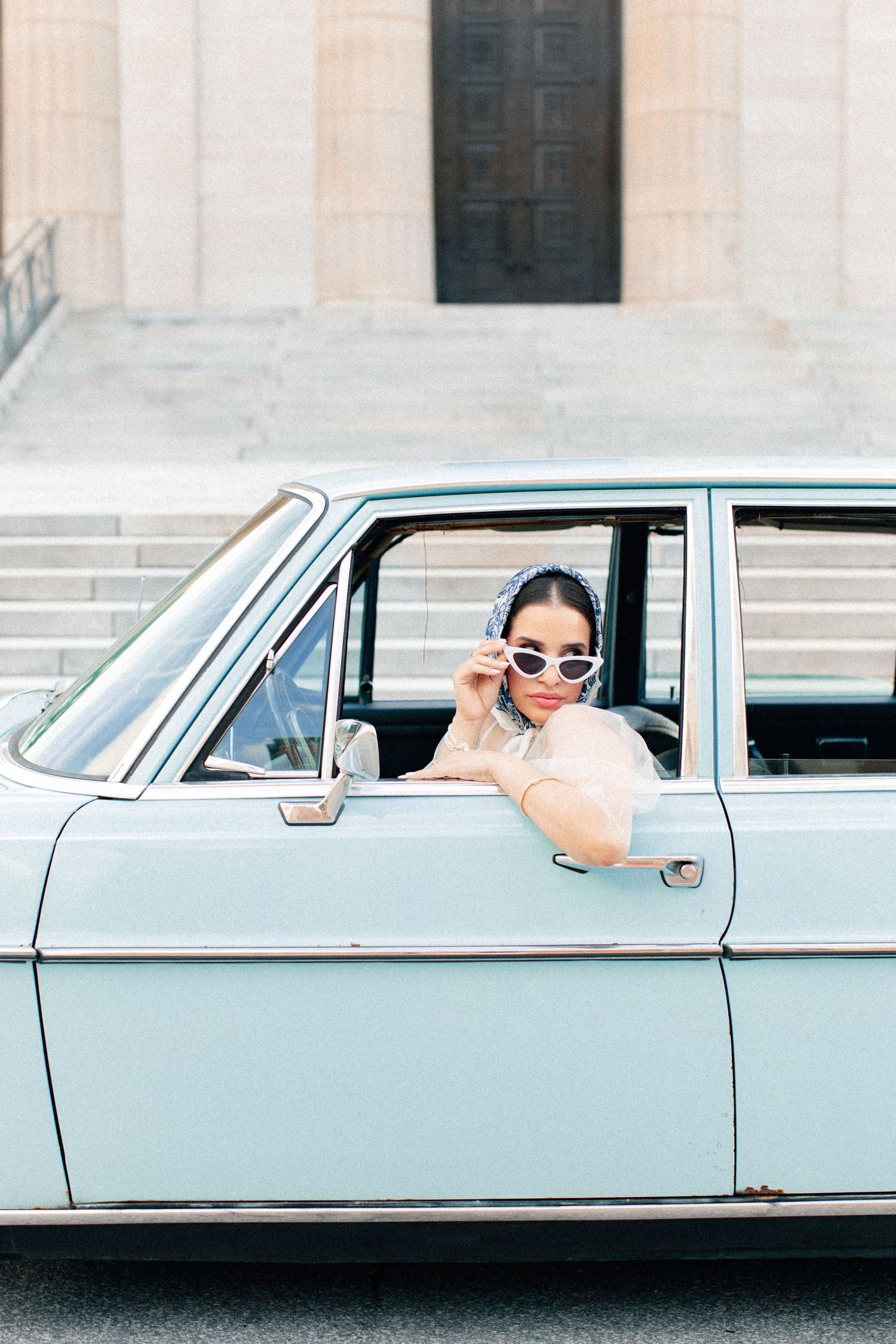 A stylish woman in sunglasses and headscarf leans out of a vintage light blue classic car parked near stone steps.