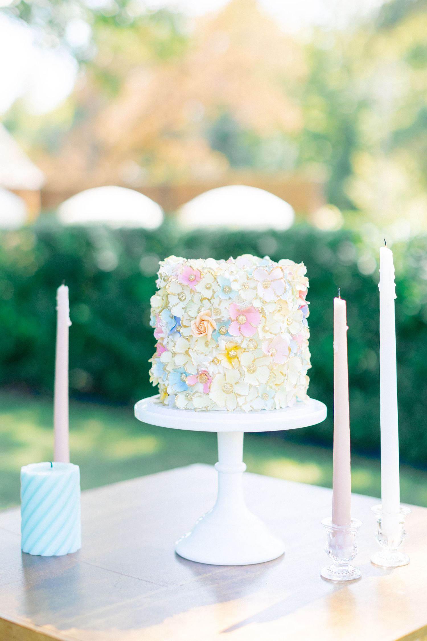 Elegant single-tier wedding cake with pastel floral decorations on white cake stand flanked by taper candles outdoors.
