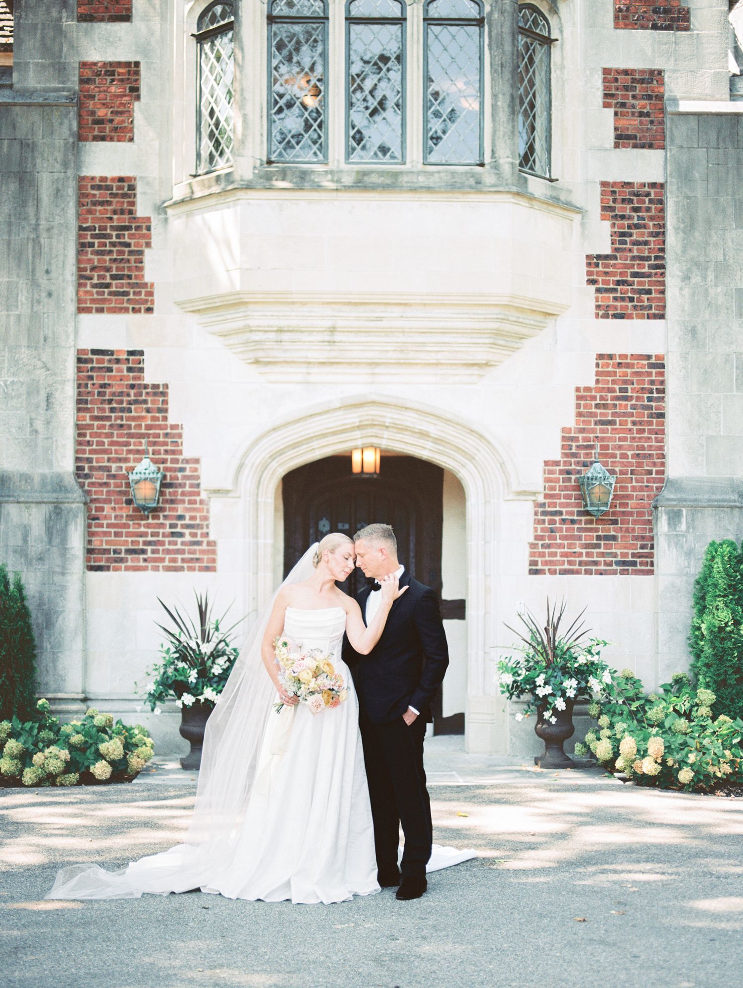 Bride and groom share a romantic moment outside an elegant brick castle venue surrounded by lush greenery on their wedding day.