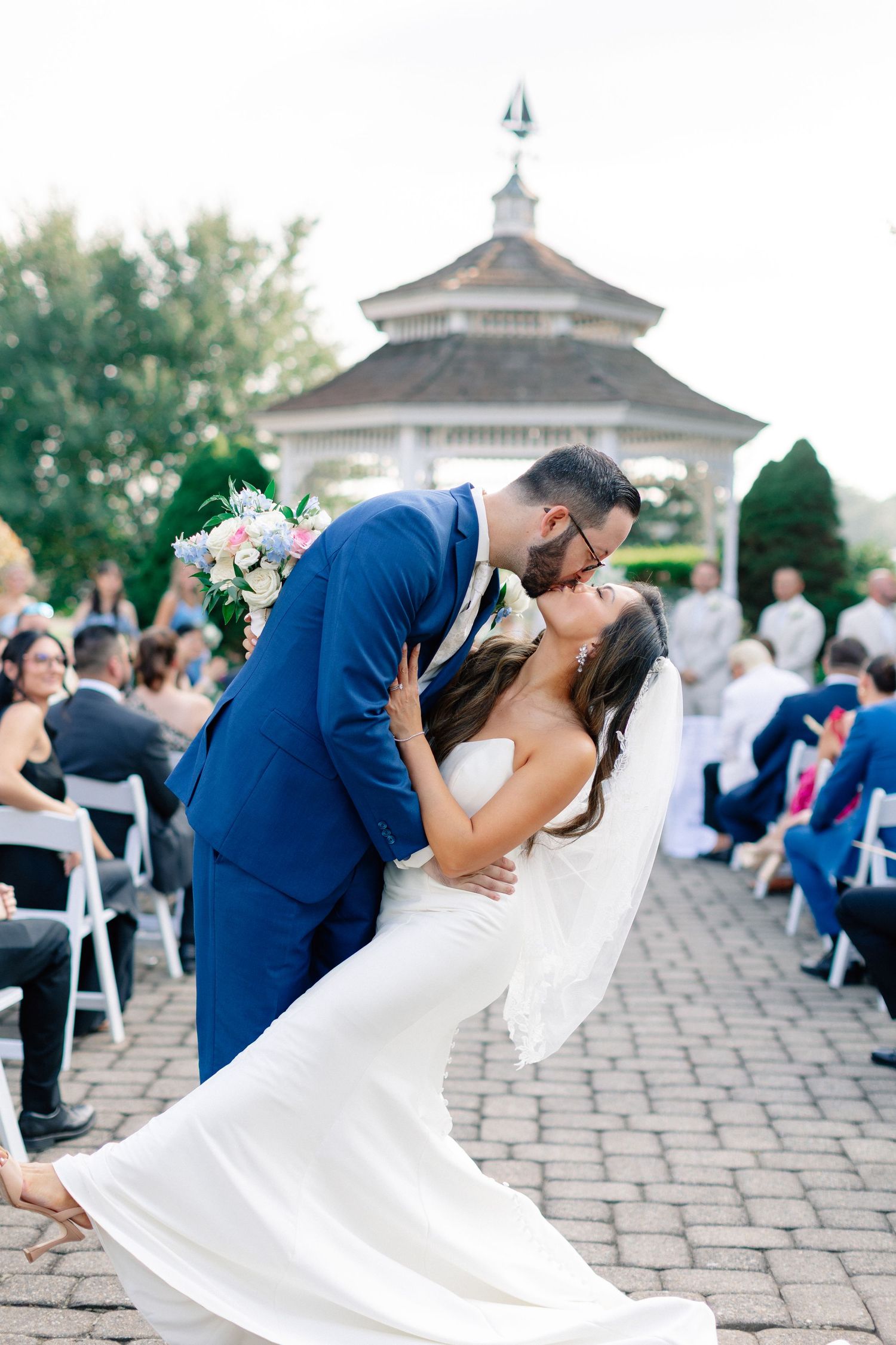 Groom in blue suit dips bride in white gown for a kiss at outdoor wedding ceremony near gazebo.
