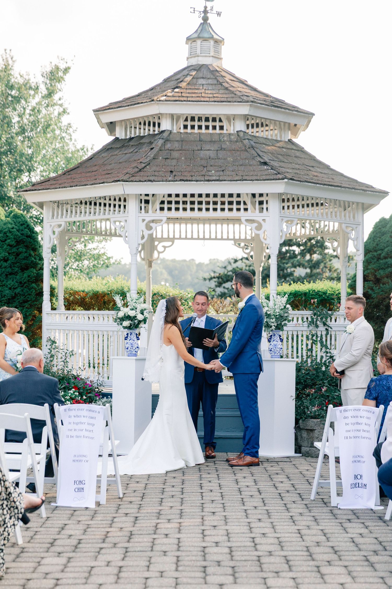 Outdoor wedding ceremony at a white gazebo with bride and groom exchanging vows, surrounded by lush greenery and white chairs.