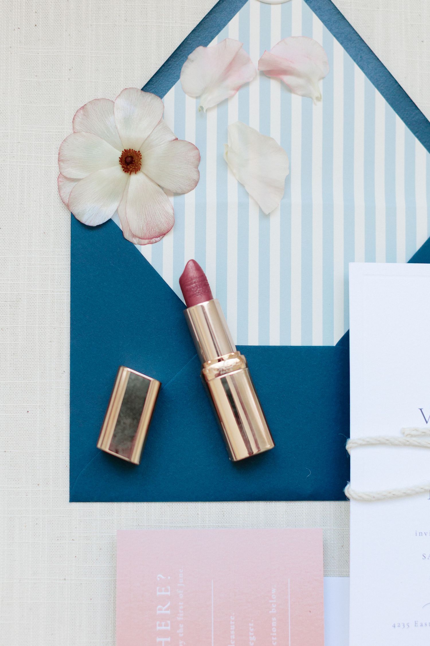 A mauve lipstick with gold casing rests on a teal envelope with white flowers and elegant wedding stationery.