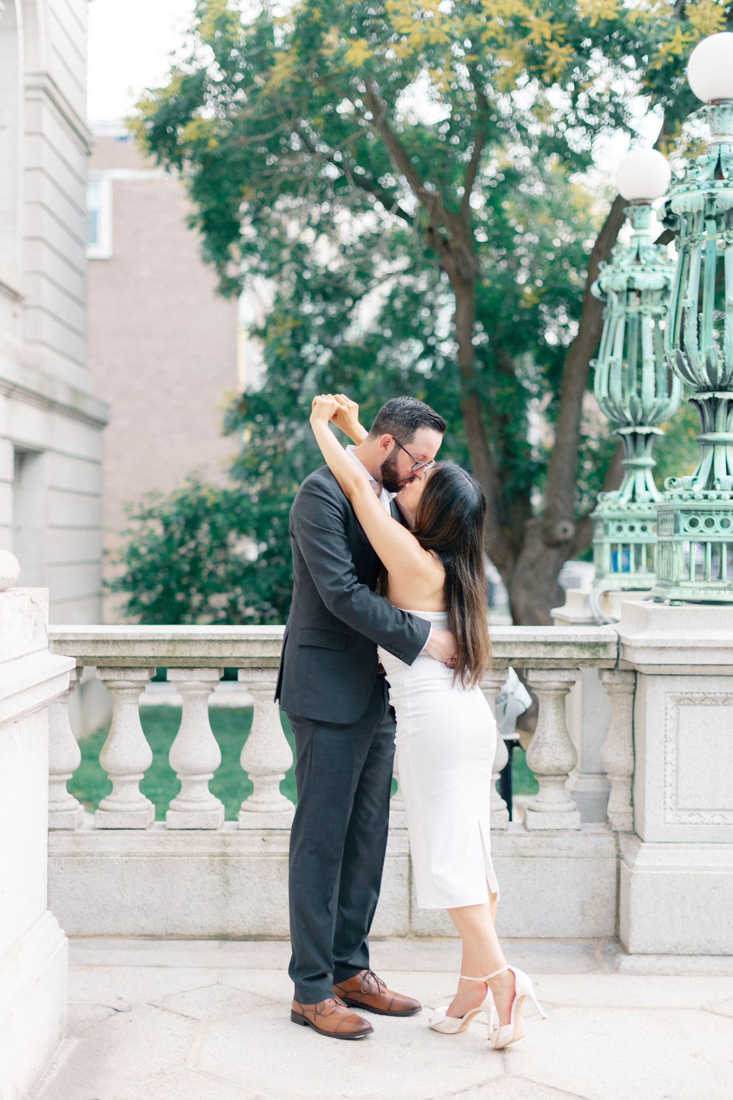 Couple sharing a kiss on stone balcony with green lamp post and trees.