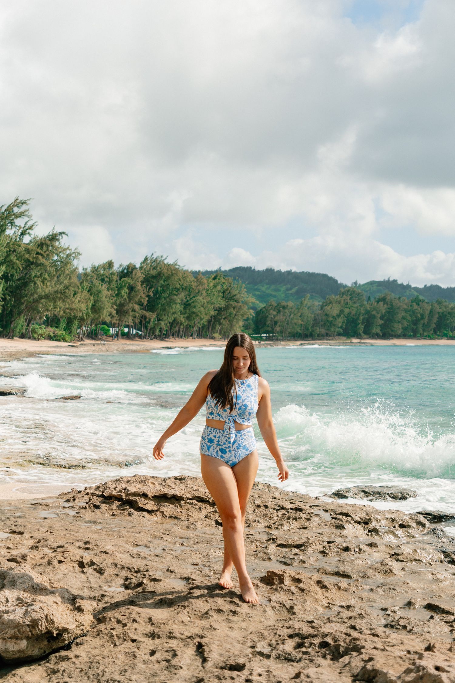 Woman in blue floral swimsuit walking on rocky Hawaiian beach with turquoise waves and lush green mountains behind.