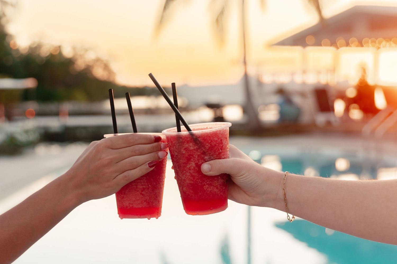 Two hands clinking watermelon slushie drinks by a tropical pool at sunset.
