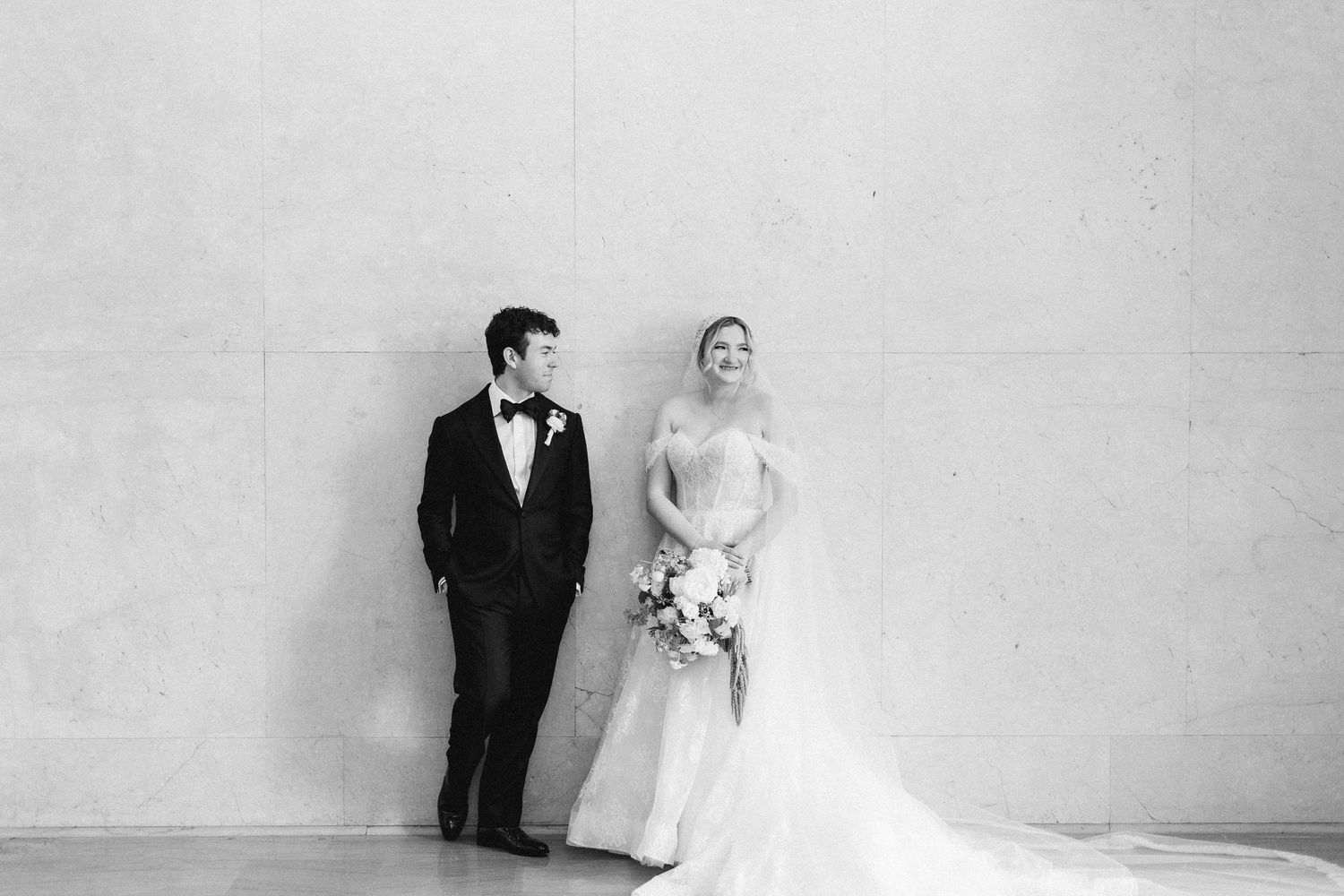 Black and white wedding photo of bride in elegant gown and groom in tuxedo standing against a wall.