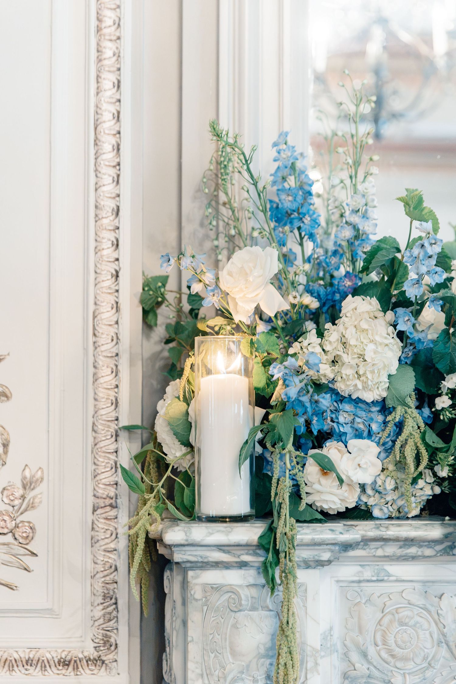 Elegant white pillar candle beside blue hydrangeas and white roses on a marble mantle in a classic interior.