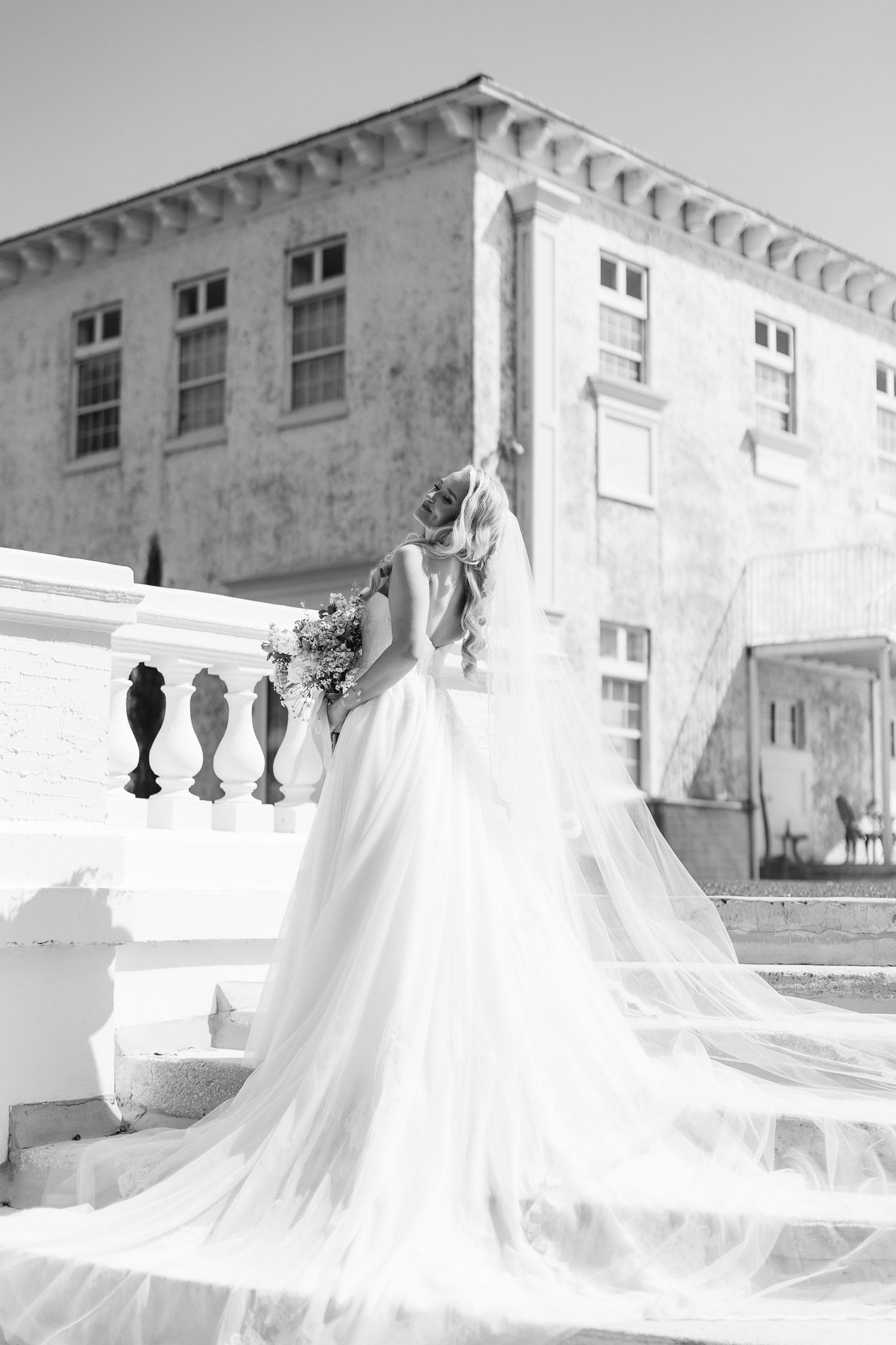 Black and white photo of a bride in a flowing gown and veil holding a bouquet on elegant white steps, wedding at Bella Cosa