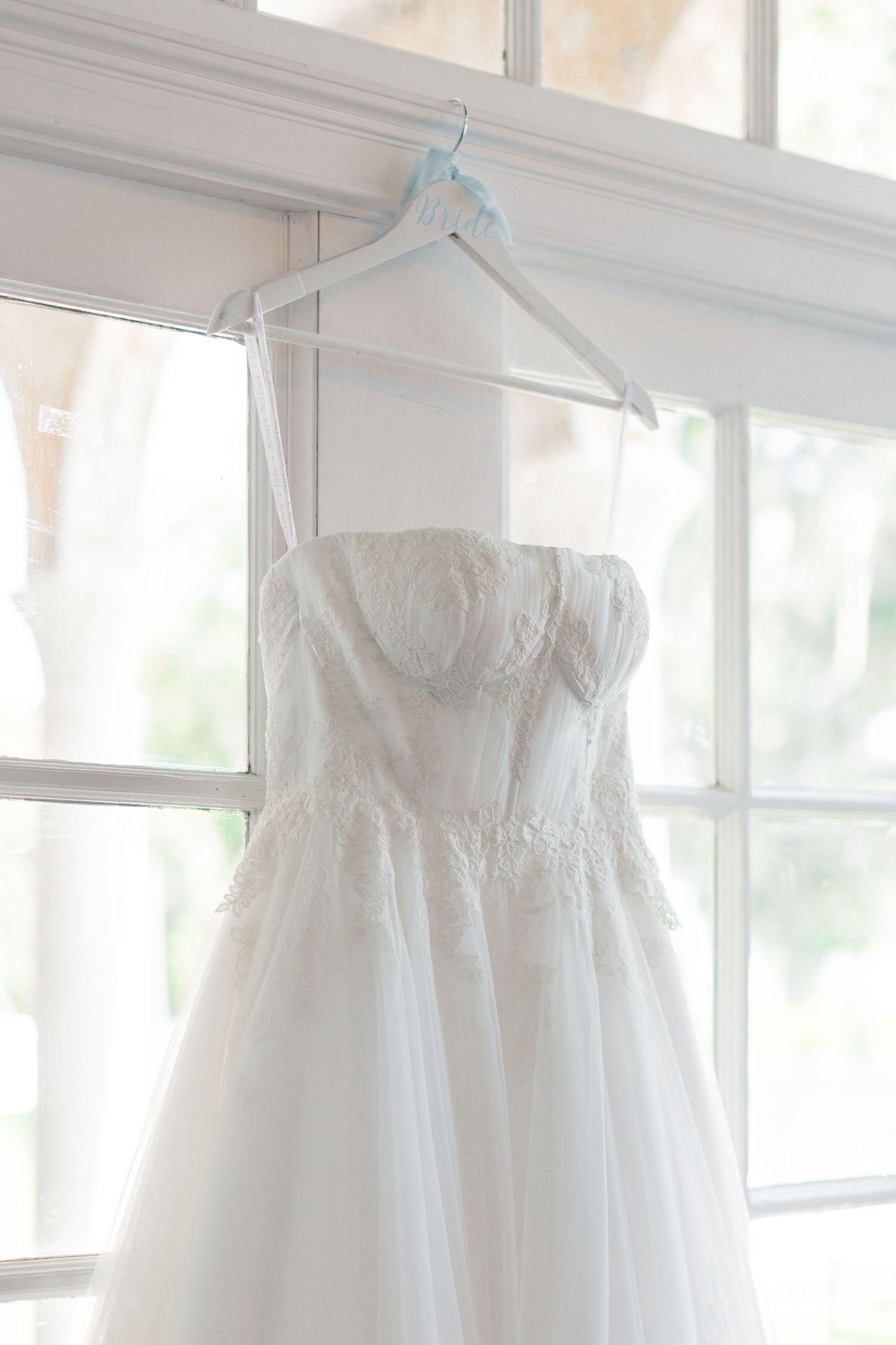 White strapless lace wedding dress hanging on a hanger in front of a bright window with natural light, wedding at Bella Cosa