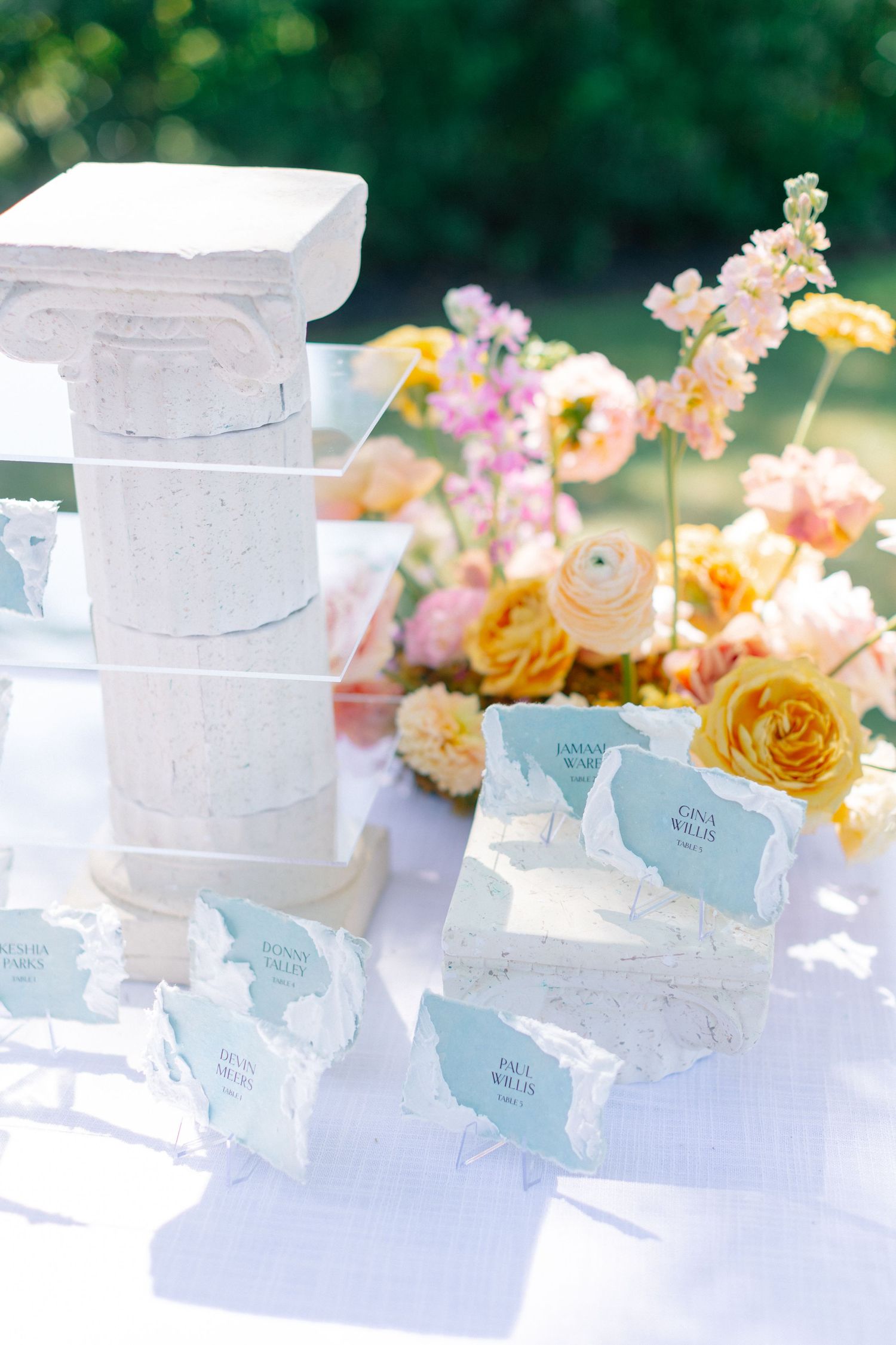 Elegant outdoor wedding escort card display with white stone column, blue place cards, and colorful floral arrangement.