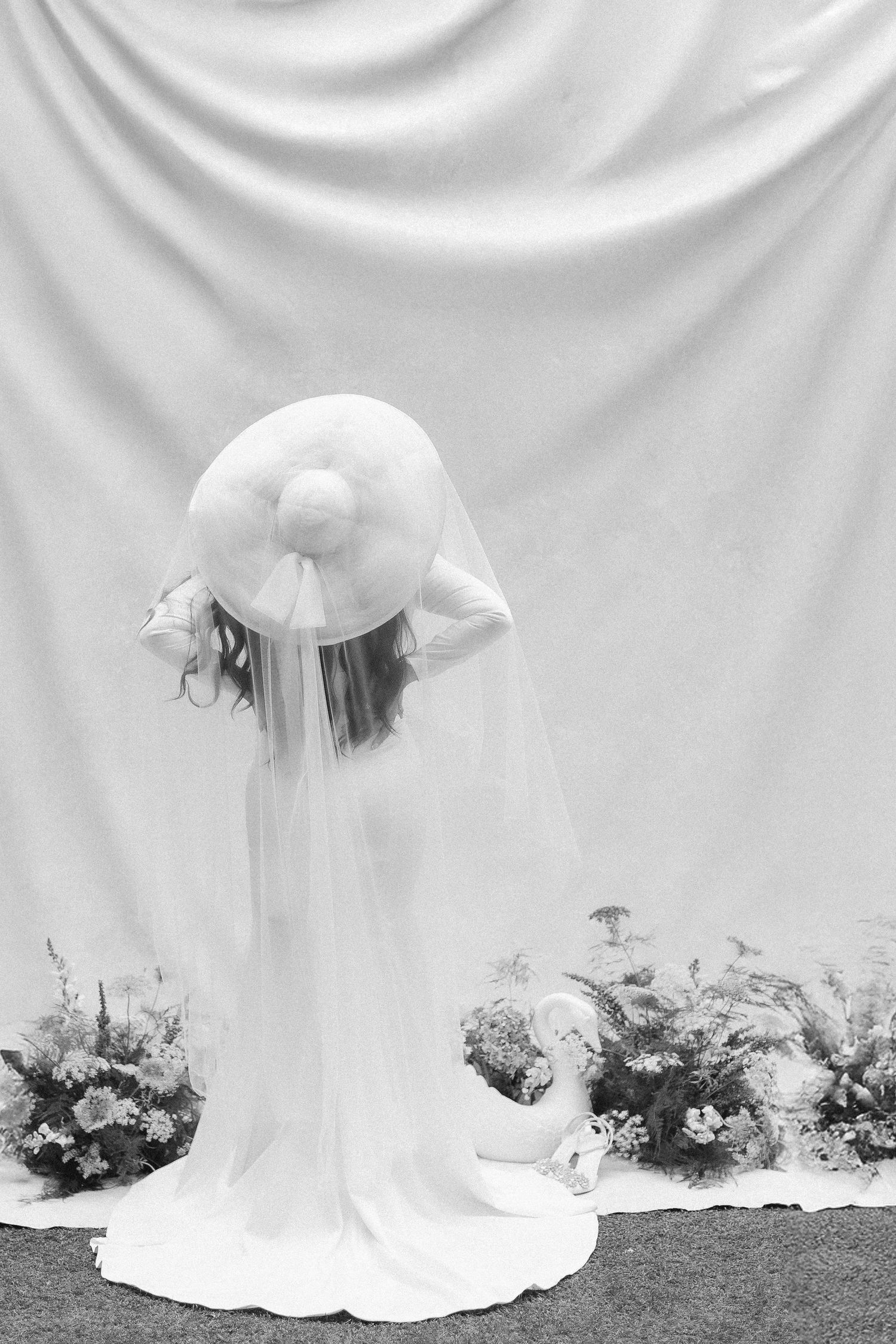 Bride in white gown and wide-brim hat with veil stands before draped fabric backdrop with floral arrangements.